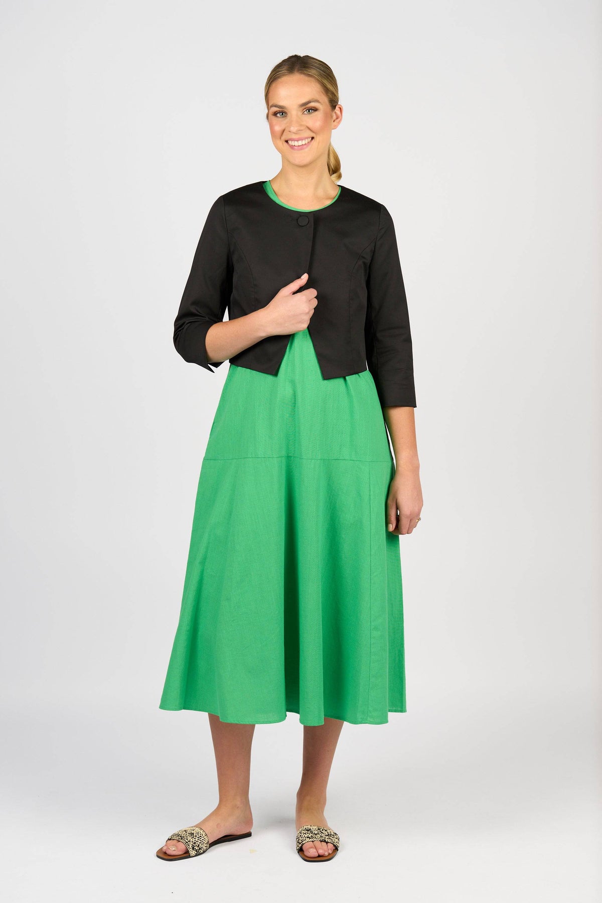 A woman smiles, wearing a Vassalli Cropped Jacket in black over a bright green midi dress and animal print flat sandals. Her blonde hair is pulled back, and she poses against a plain white background.