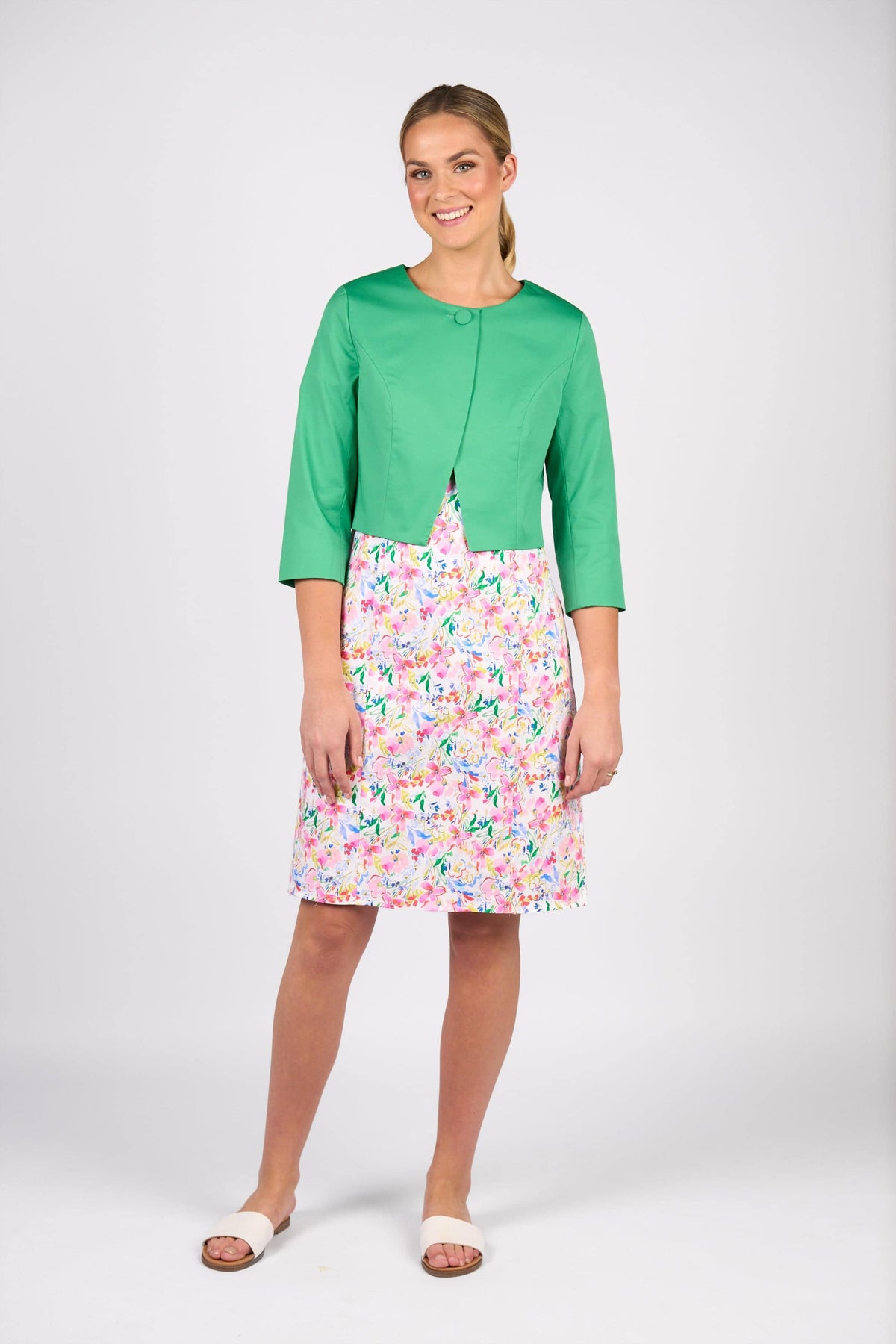 A woman smiles in a Vassalli Cropped Jacket - Clover, paired with a knee-length white floral dress and white open-toe shoes, standing against a plain light gray background.