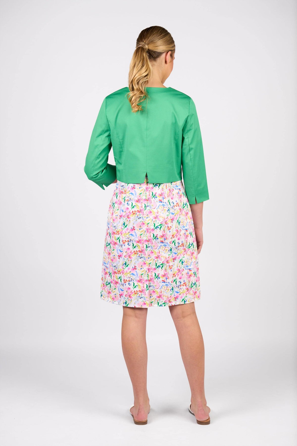 A woman faces away, wearing the Vassalli Cropped Jacket - Clover over a colorful floral knee-length dress. Her blonde hair is tied back and she pairs nude slip-on shoes with the outfit, set against a plain white background.