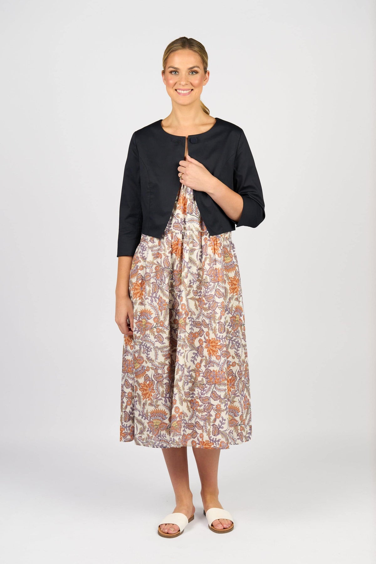 A woman smiles, wearing the Vassalli Cropped Jacket - Ink over a floral, earthy-toned mid-length dress. Her light hair is tied back, and she pairs the look with white open-toe sandals against a plain light background.