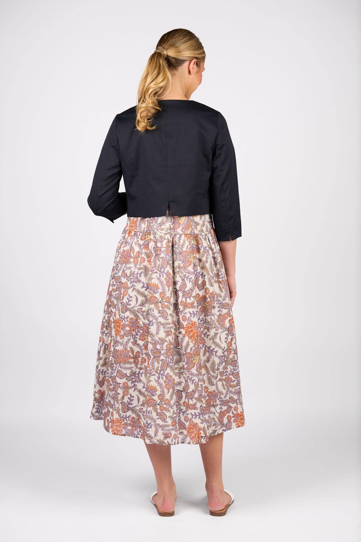 A barefoot woman with blonde hair in a low ponytail, seen from behind on a white background, wears Vassalli's Cropped Jacket - Ink over a muted orange, purple, and cream floral-patterned calf-length skirt.