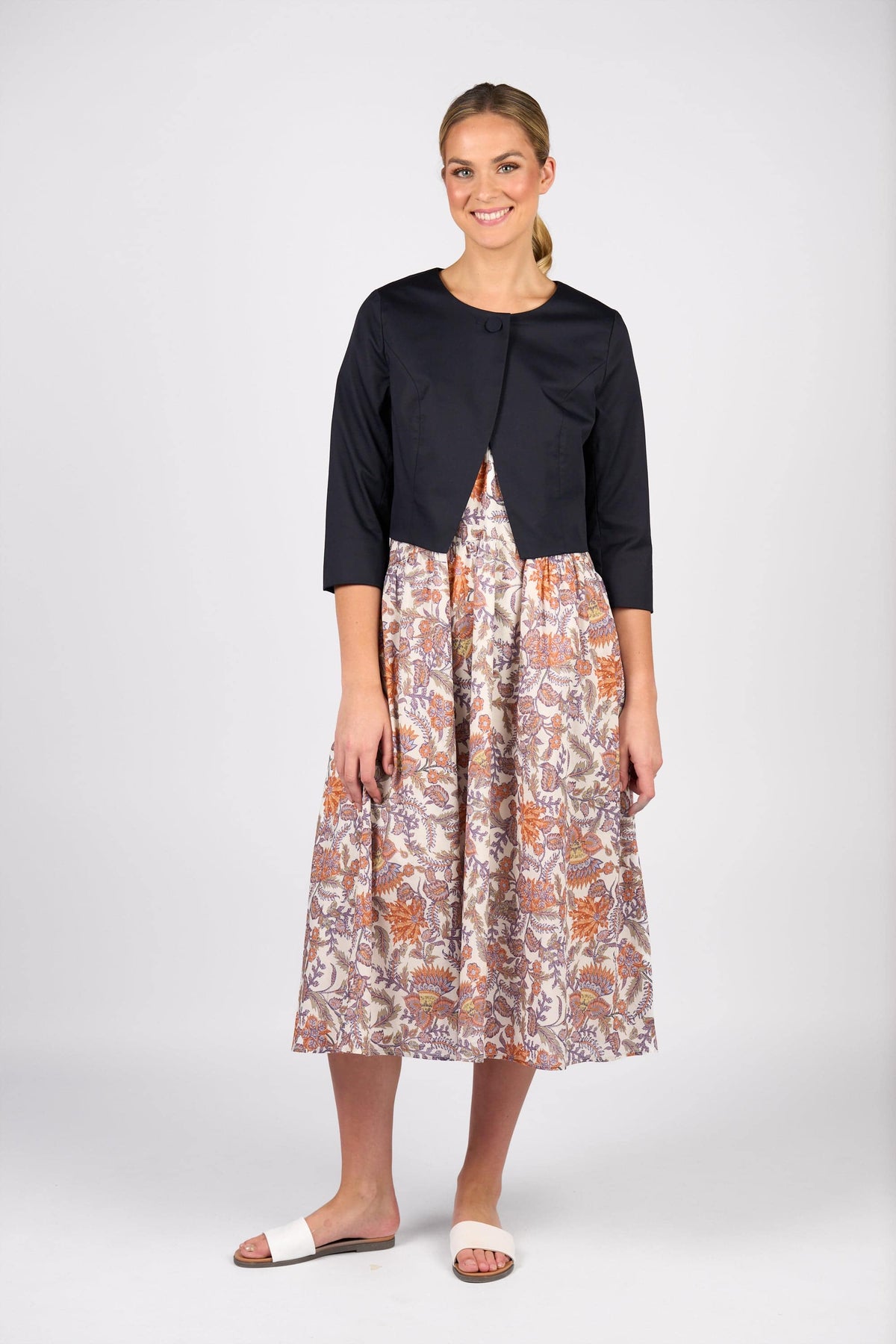 A woman smiles in a Vassalli Cropped Jacket - Ink, layered over a floral knee-length dress. She wears light slide sandals, her hair tied back, against a plain white background.