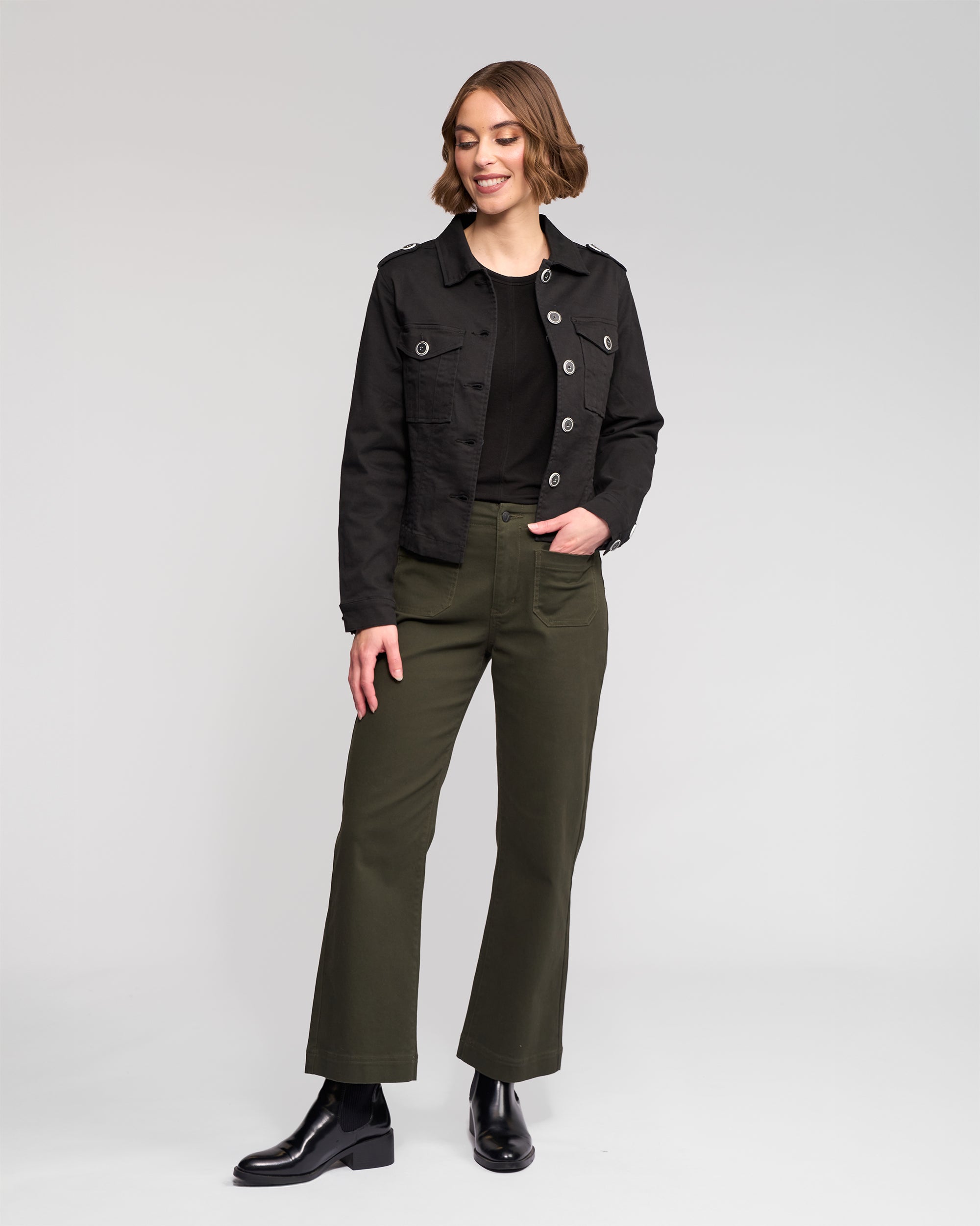 A woman with short brown hair wears the Vassalli Satellite Jacket in black over a black top, olive green pants, and black boots. She stands slightly to the side with one hand in her pocket against a light gray background.
