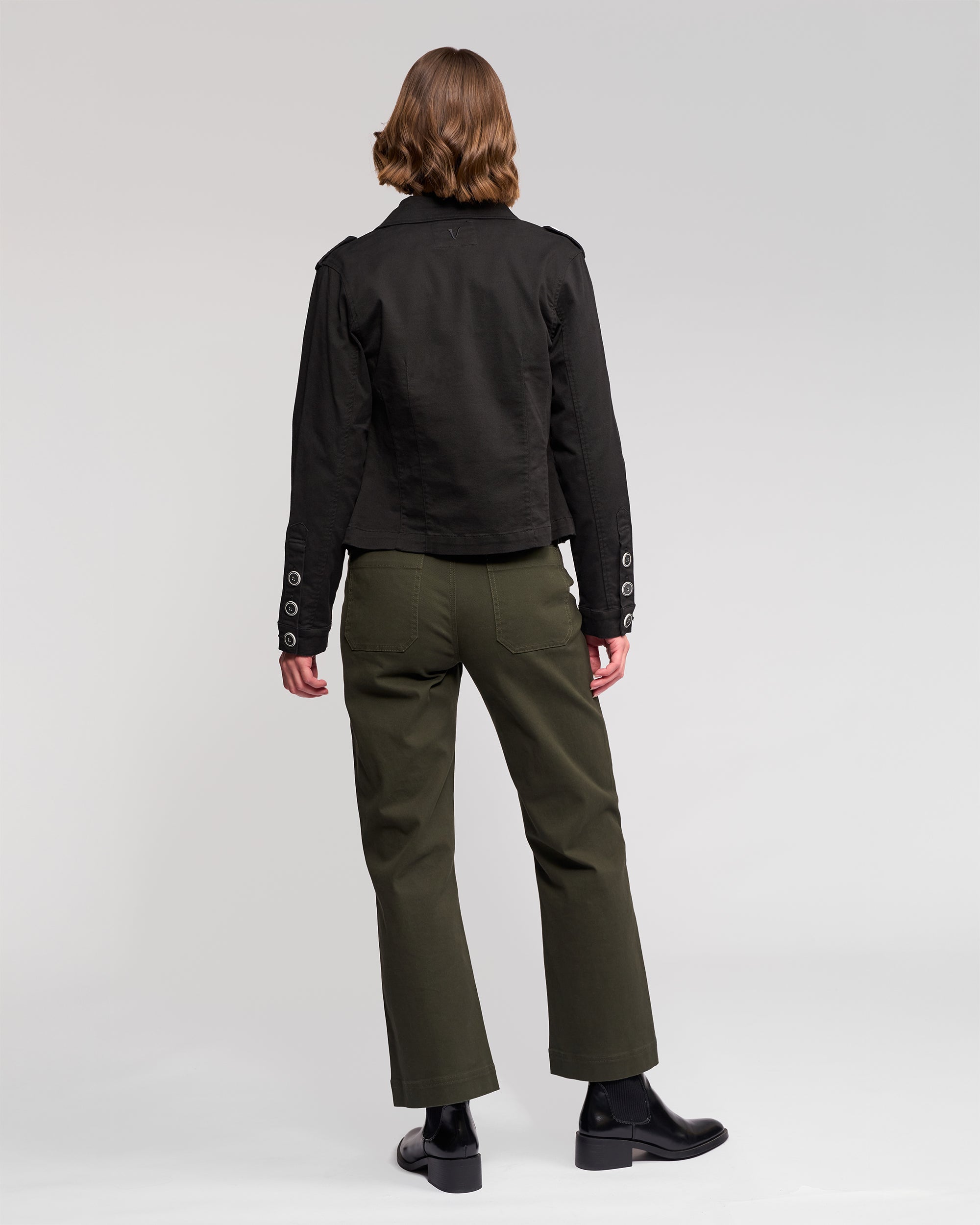 A person with short, wavy hair faces away, wearing the Vassalli Satellite Jacket in black, olive green pants, and black ankle boots against a plain light background.