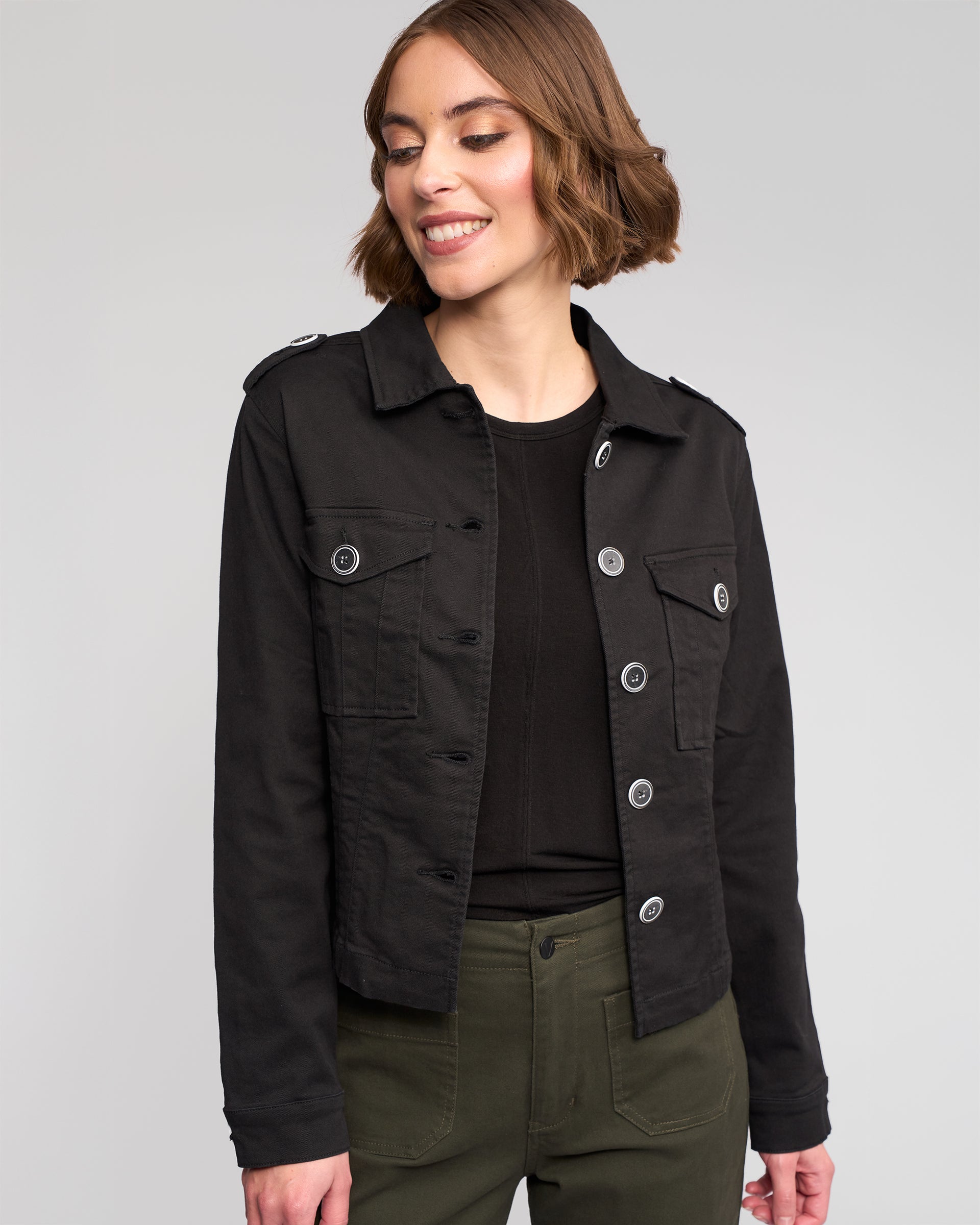 A woman with short brown hair smiles while wearing the Vassalli Satellite Jacket in black over a black top and olive green pants, standing against a plain light gray background.