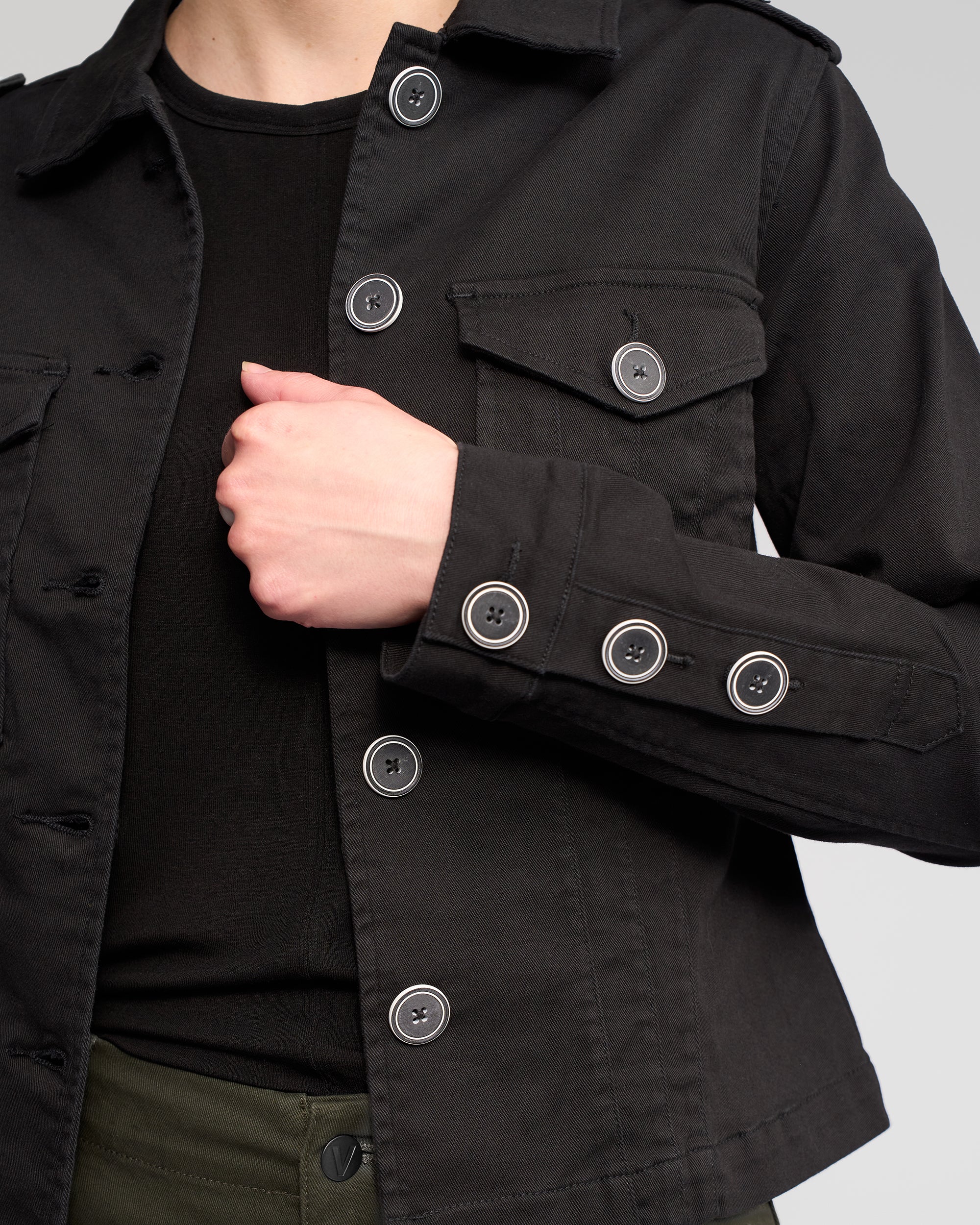 A person wears the Vassalli Satellite Jacket in black over a black shirt and olive green pants, adjusting the jacket near the chest. The image is cropped to display only the torso and part of one arm.