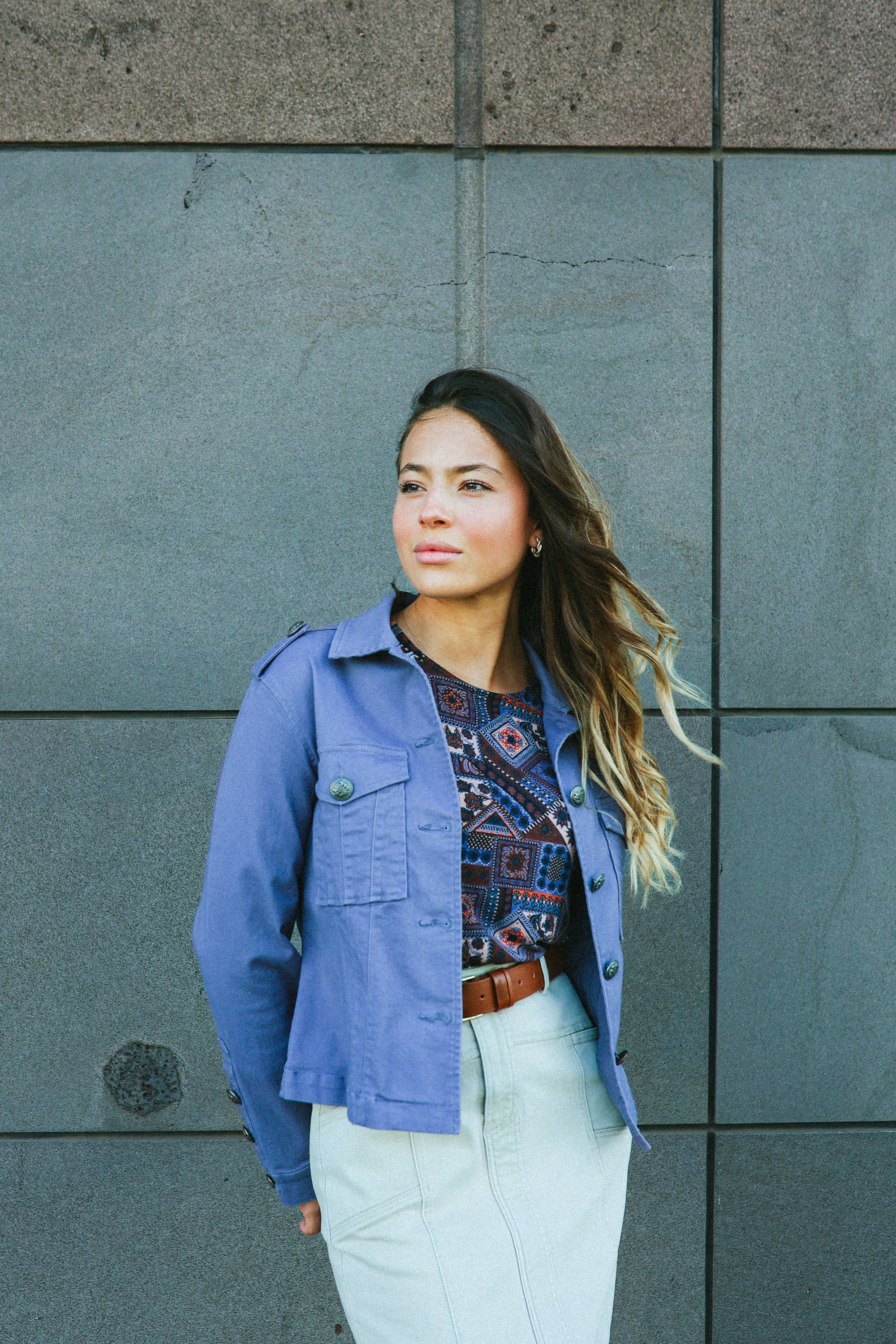A woman with long, wavy hair stands before a gray tiled wall, wearing the Vassalli Satellite Jacket in Denim Blue over a patterned top and light skirt, looking confidently to the side.