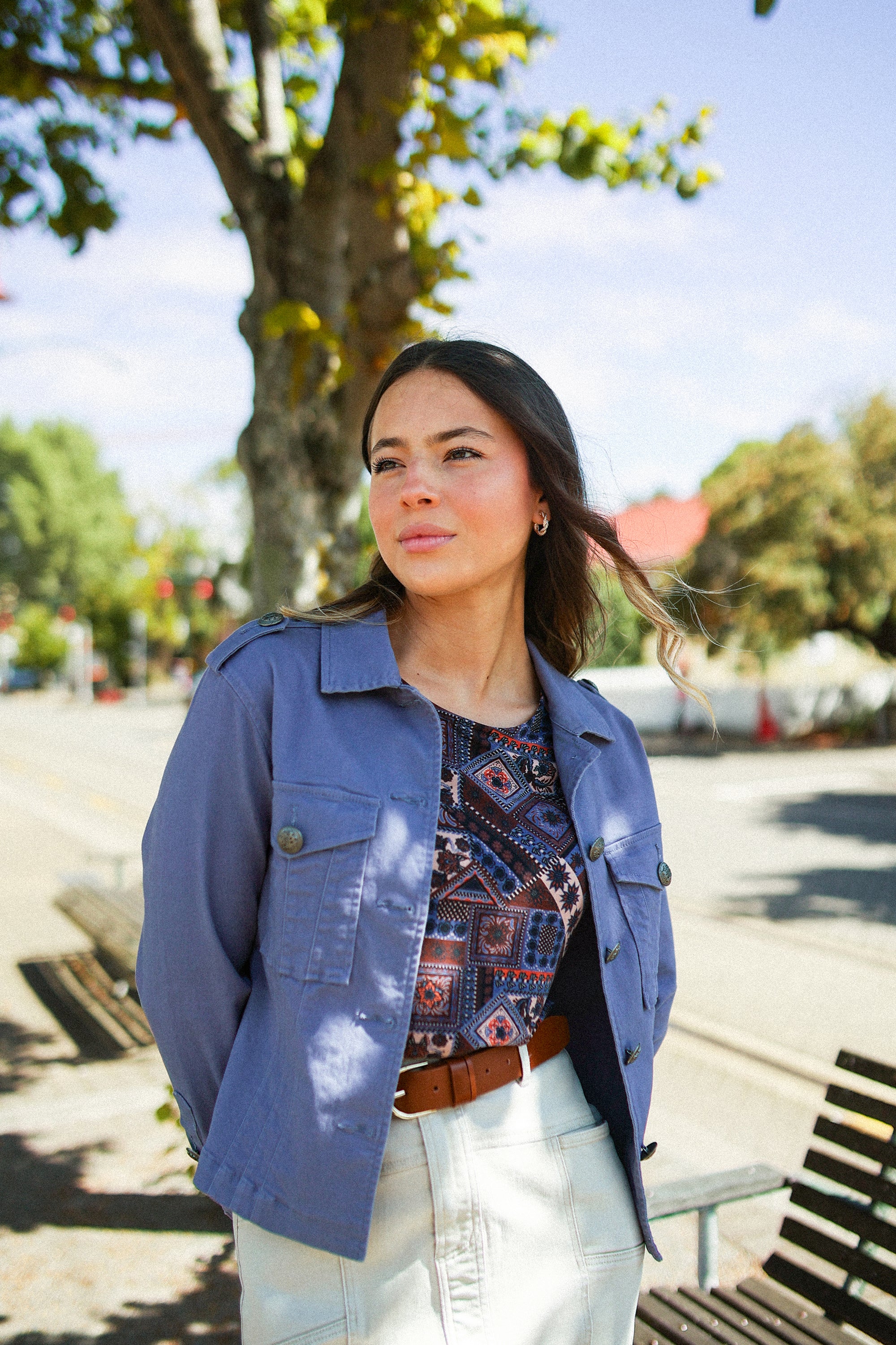 A woman with long dark hair stands outdoors on a sunny day, wearing the Vassalli Satellite Jacket in Denim Blue over a patterned top and white skirt. She gazes off to the side, with trees and a street in the background.