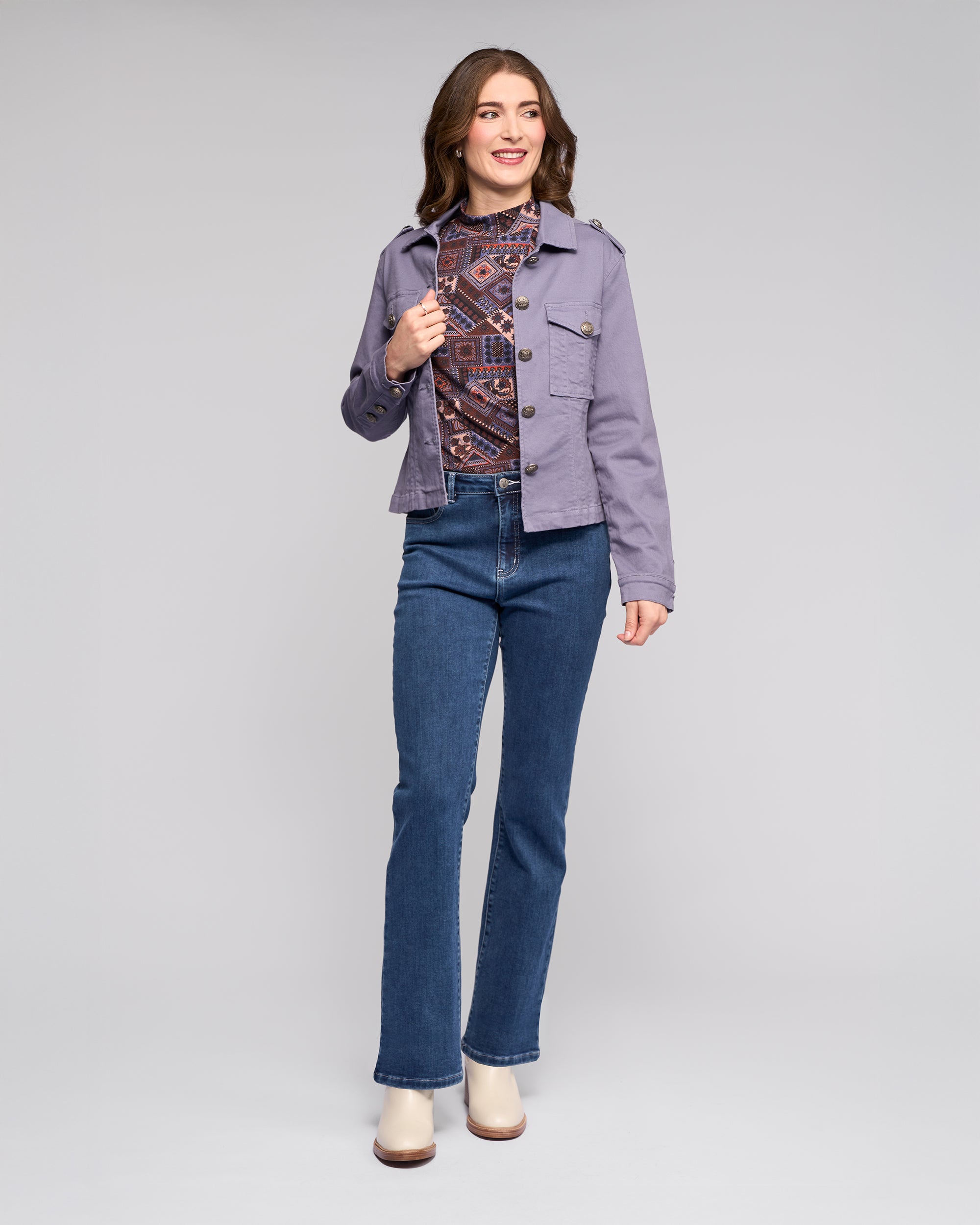 A woman smiles, wearing the Vassalli Satellite Jacket in Denim Blue over a patterned top, blue jeans, and cream boots, standing against a plain light gray background.