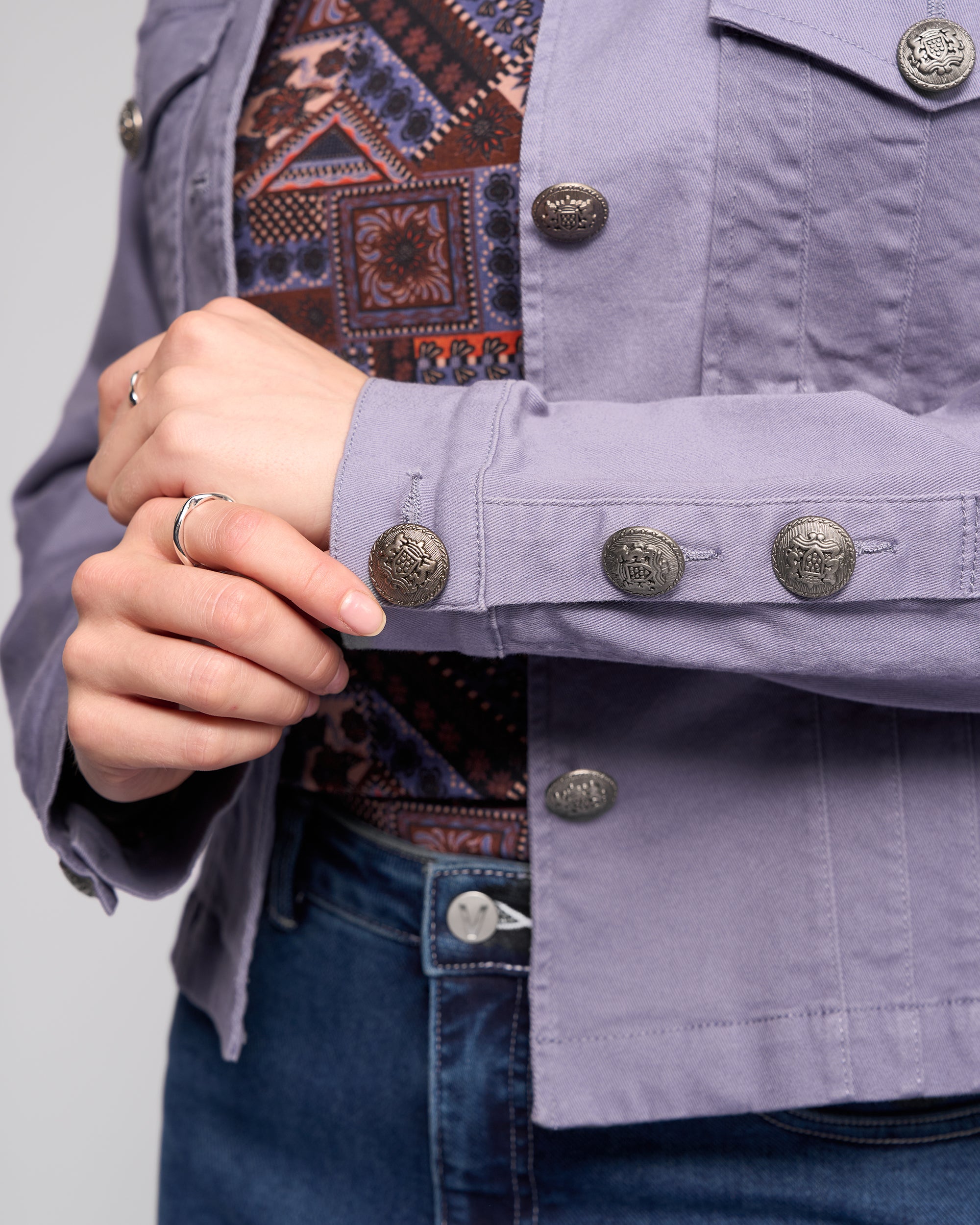 A person in a patterned top, blue jeans, and the Vassalli Satellite Jacket - Denim Blue adjusts their sleeve cuff, accessorized with several thin rings on one hand.