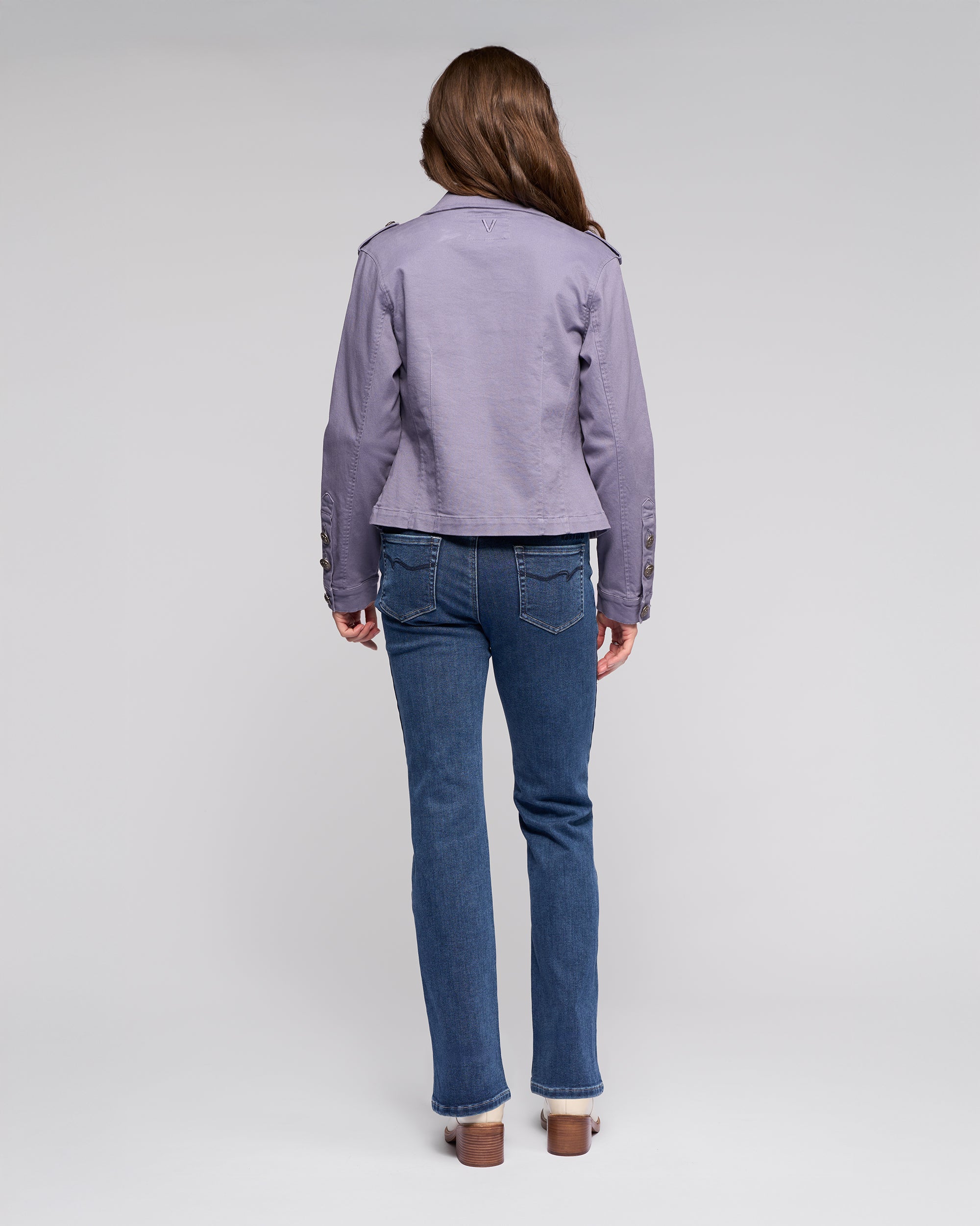 A person with long brown hair faces away, wearing the Vassalli Satellite Jacket in Denim Blue, paired with blue jeans and heeled shoes, set against a plain light gray background.