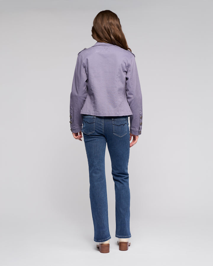 A person with long brown hair faces away, wearing the Vassalli Satellite Jacket in Denim Blue, paired with blue jeans and heeled shoes, set against a plain light gray background.