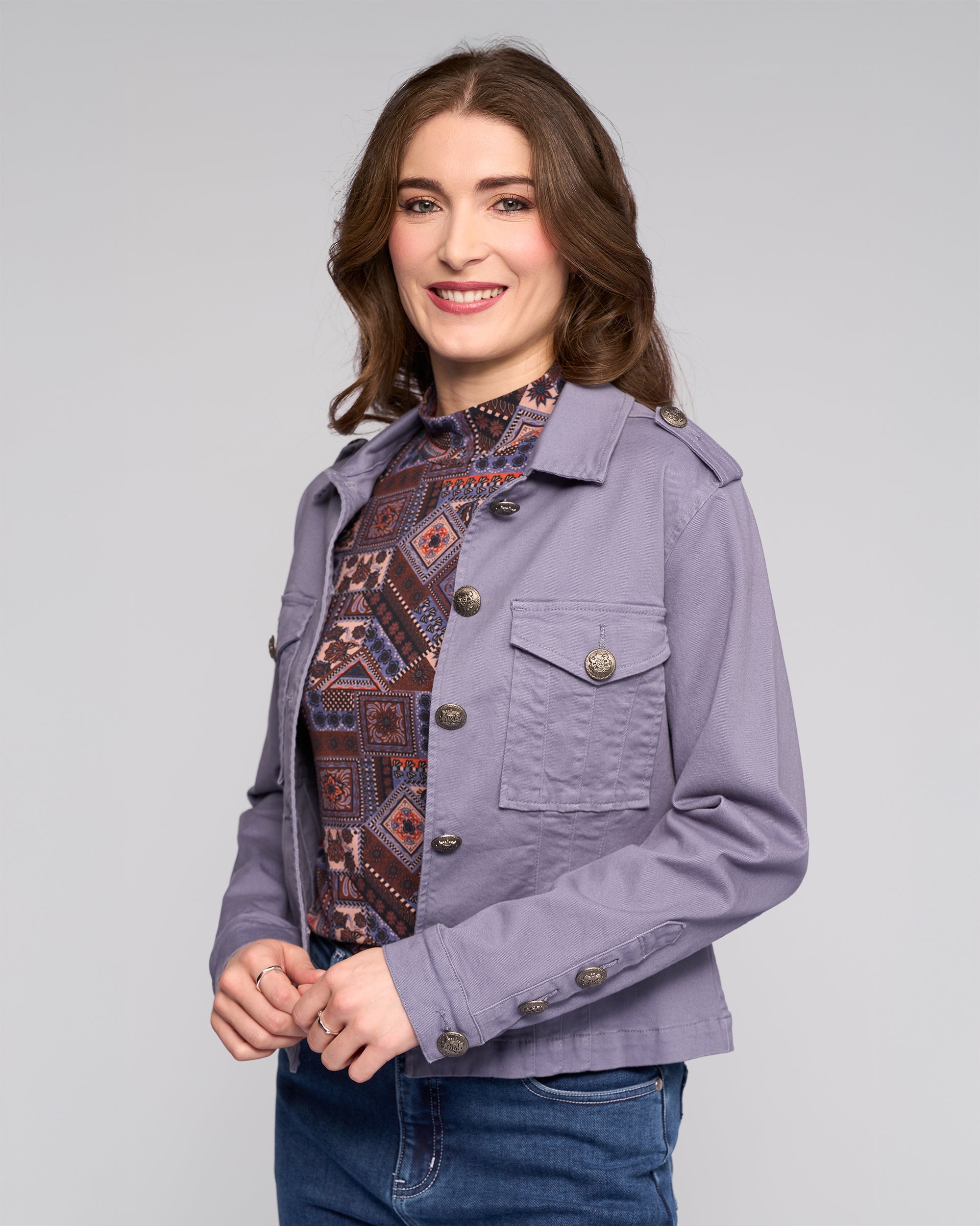 A woman with long brown hair smiles while wearing the Vassalli Satellite Jacket in Denim Blue over a patterned shirt and blue jeans, standing against a light gray background.