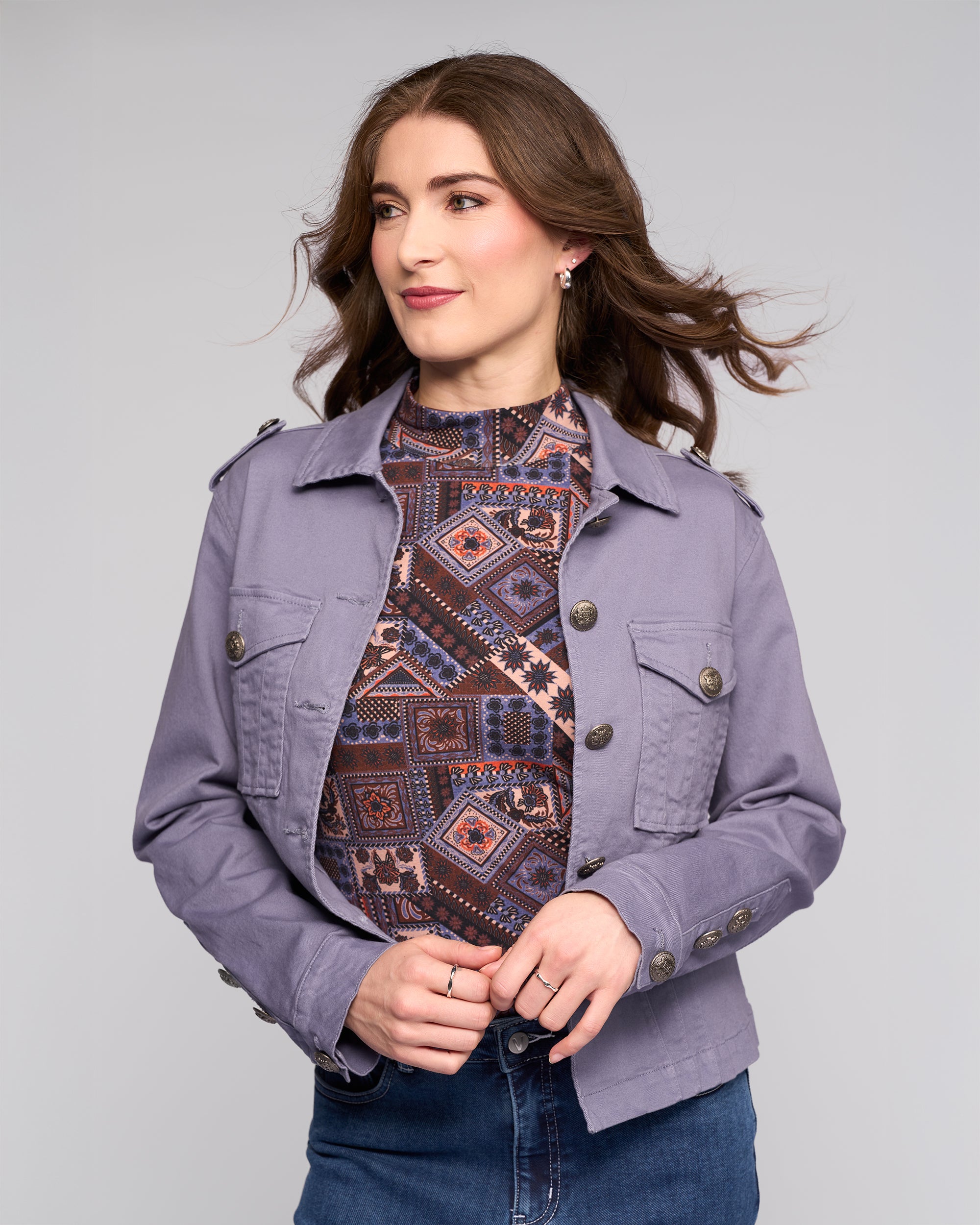A woman with wavy brown hair wears the Vassalli Satellite Jacket in Denim Blue over a patterned high-neck top and blue jeans, standing against a light gray background and looking slightly to the side with a subtle smile.