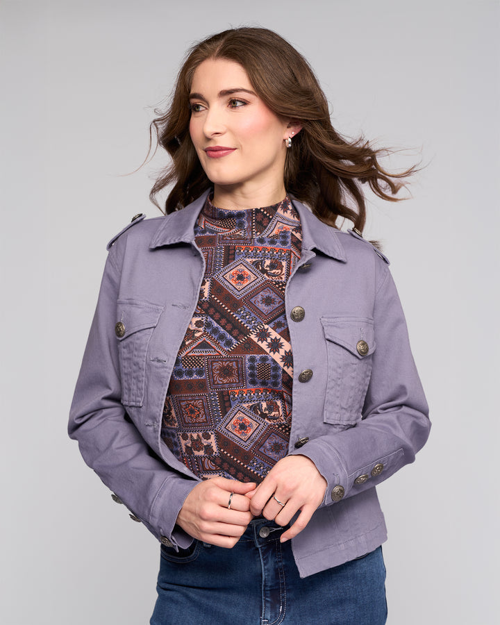 A woman with wavy brown hair wears the Vassalli Satellite Jacket in Denim Blue over a patterned high-neck top and blue jeans, standing against a light gray background and looking slightly to the side with a subtle smile.