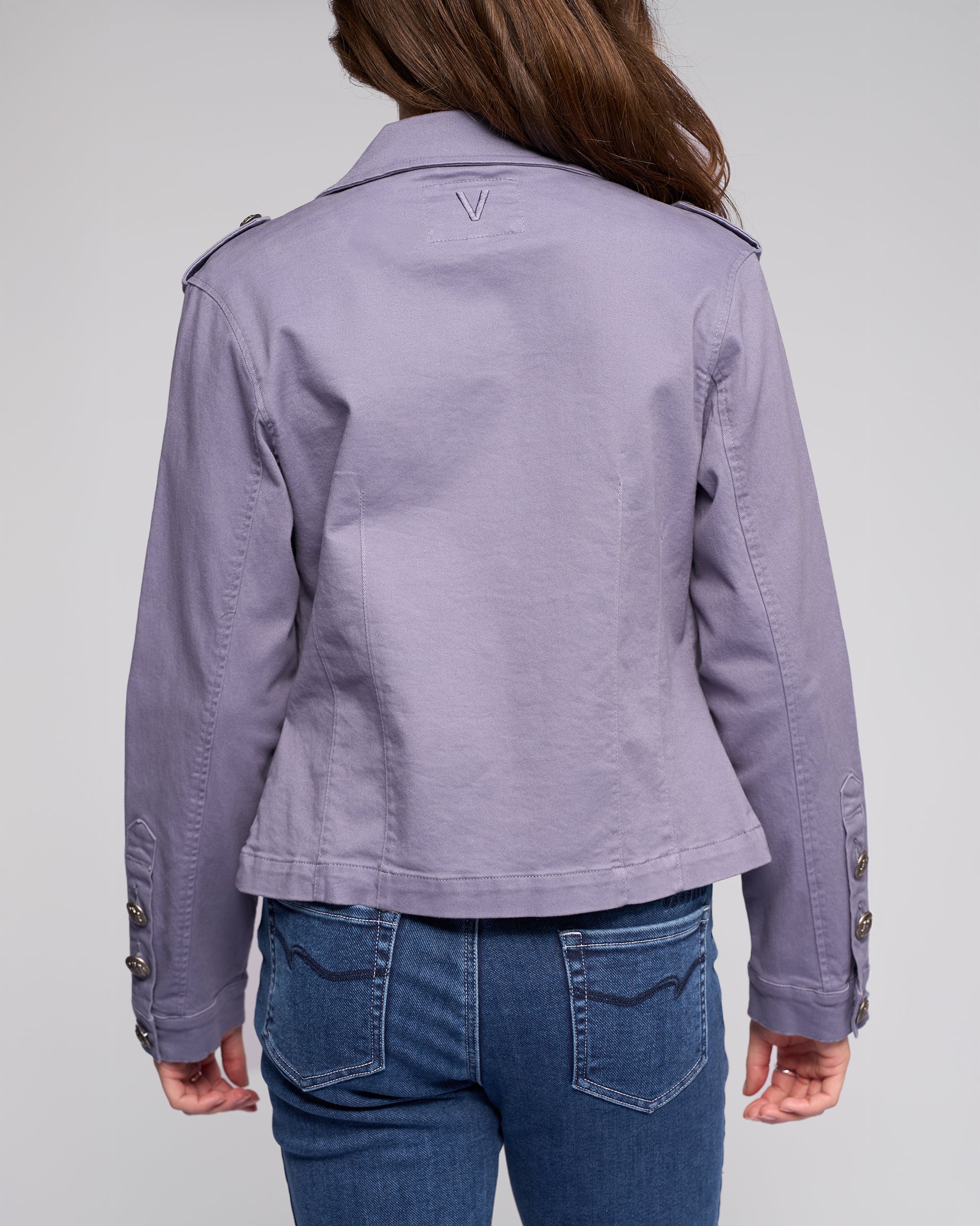 A person with long brown hair is seen from behind, wearing the Vassalli Satellite Jacket in Denim Blue and blue jeans. The slim-fit jacket features button details on the sleeves, set against a plain light gray background.