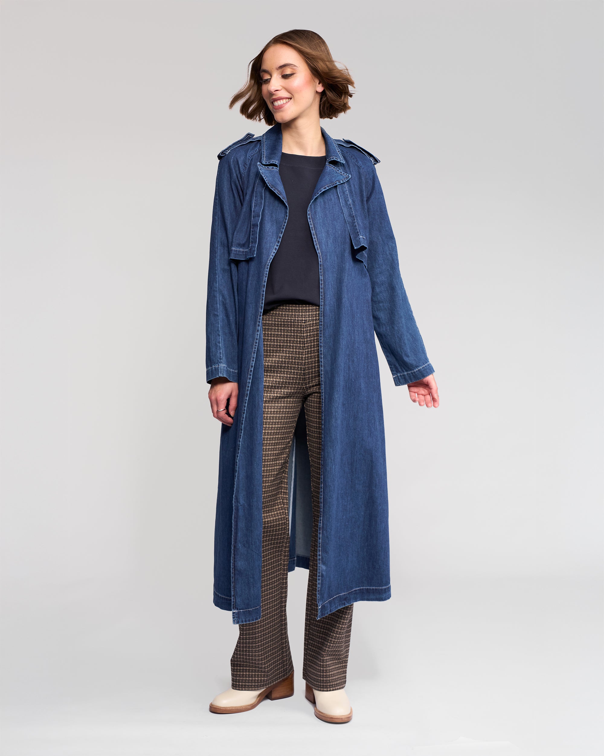 A woman smiles, wearing the Vassalli Galaxy Trench - Dark Blue over a black top and brown checkered pants with beige shoes, standing against a plain light gray background.