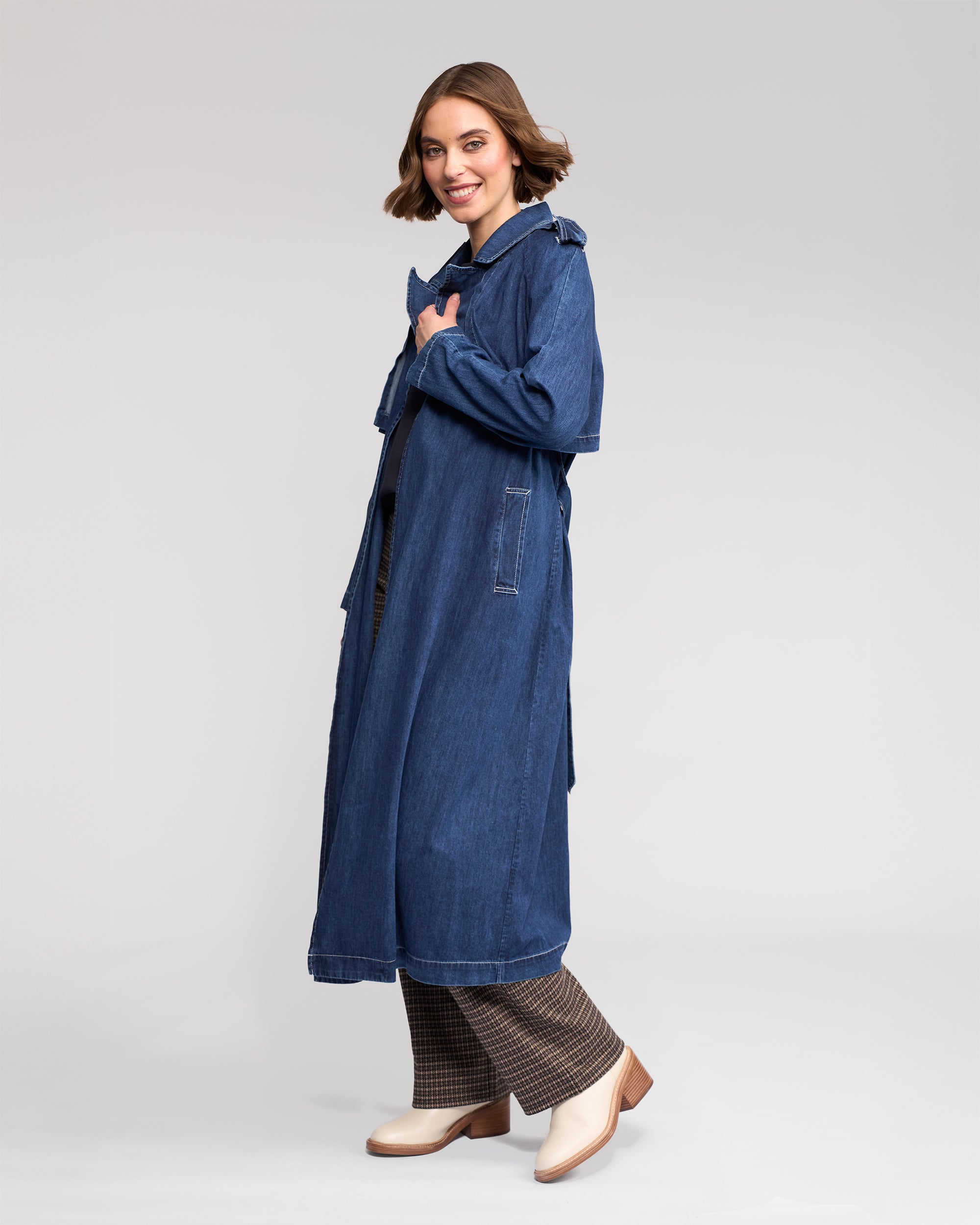 A woman smiles in the Vassalli Galaxy Trench - Dark Blue, a relaxed-fit denim coat with classic trench detailing, styled over plaid pants and cream ankle boots, set against a plain light gray background.