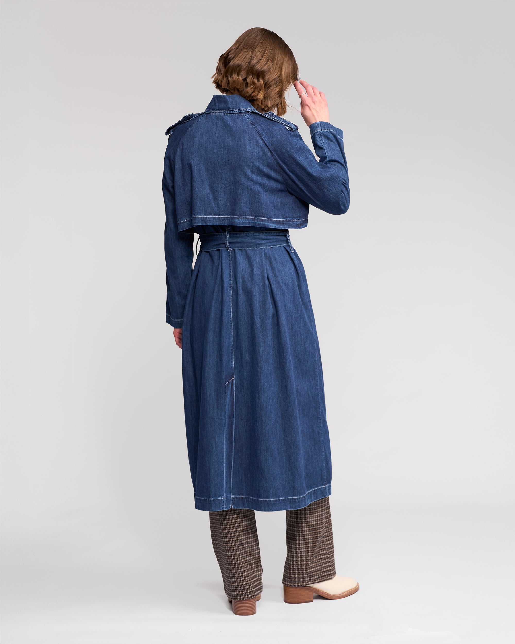 A person stands facing away, wearing the Vassalli Galaxy Trench in dark blue over brown checkered pants and beige shoes, with a relaxed posture, head slightly turned and one hand near their face, against a plain light-colored background.