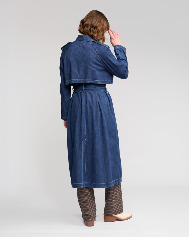 A person stands facing away, wearing the Vassalli Galaxy Trench in dark blue over brown checkered pants and beige shoes, with a relaxed posture, head slightly turned and one hand near their face, against a plain light-colored background.