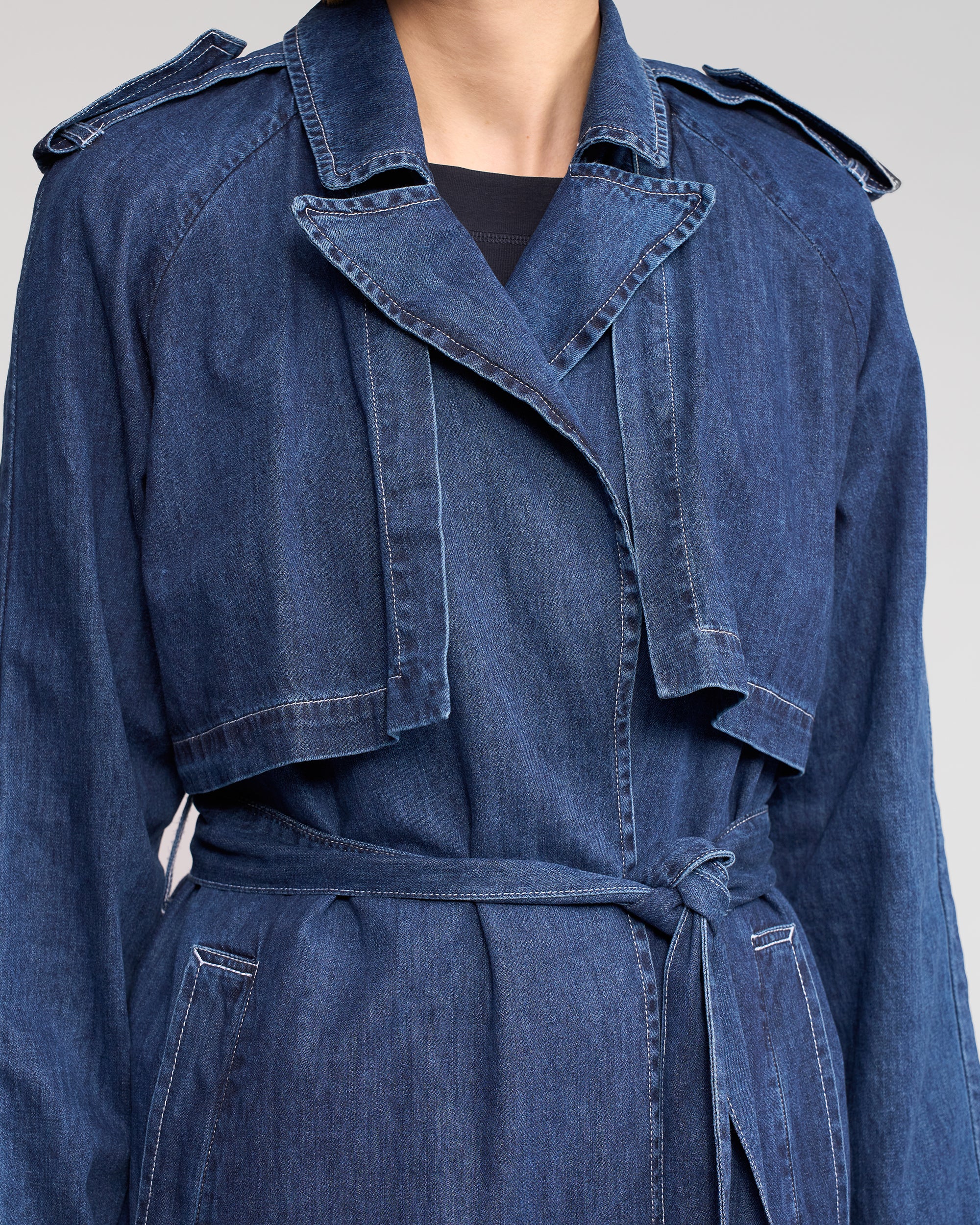 A person in the Vassalli Galaxy Trench - Dark Blue, a relaxed-fit denim coat with a layered collar and belted waist, shown from shoulders to mid-thigh against a plain background.
