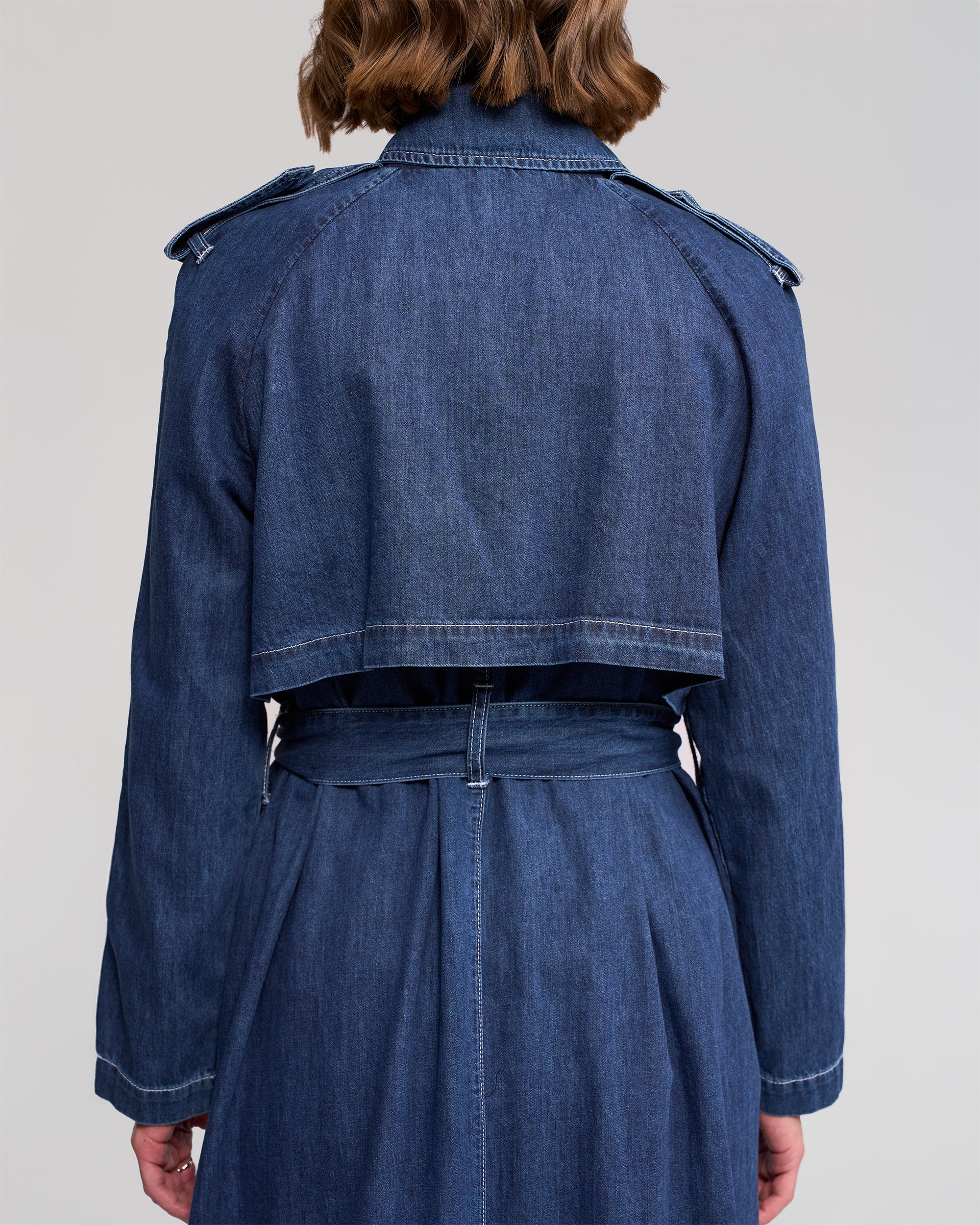 A person with shoulder-length brown hair is shown from behind wearing the Vassalli Galaxy Trench in dark blue, a long-sleeved denim coat featuring classic trench details like a cropped back flap and matching belt.
