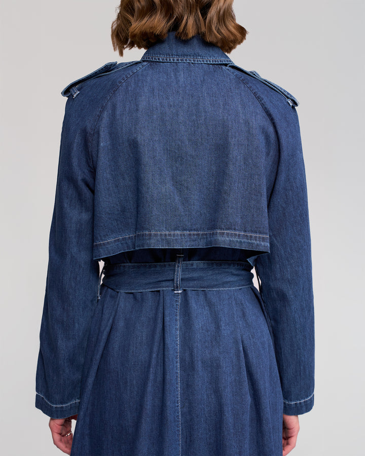 A person with shoulder-length brown hair is shown from behind wearing the Vassalli Galaxy Trench in dark blue, a long-sleeved denim coat featuring classic trench details like a cropped back flap and matching belt.