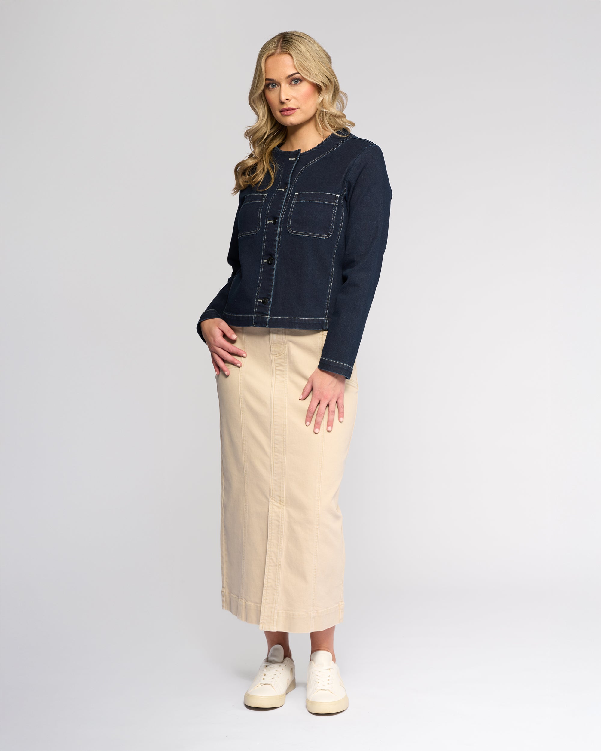 A woman with long blonde hair models the Vassalli Cable Jacket in Ink Denim, featuring front pockets and no collar, paired with a beige ankle-length skirt and white sneakers against a plain light gray background.