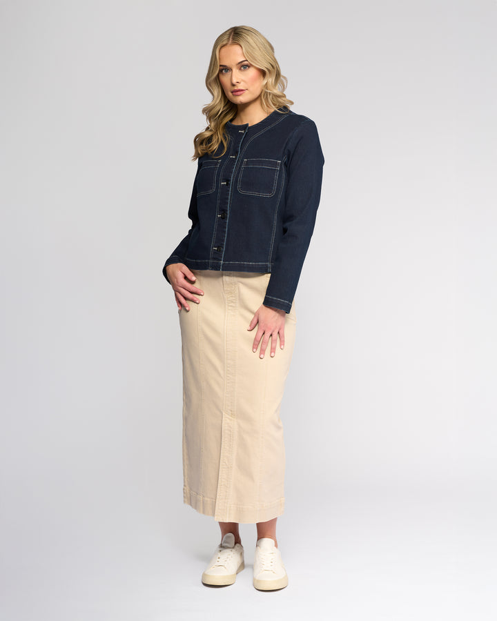 A woman with long blonde hair models the Vassalli Cable Jacket in Ink Denim, featuring front pockets and no collar, paired with a beige ankle-length skirt and white sneakers against a plain light gray background.