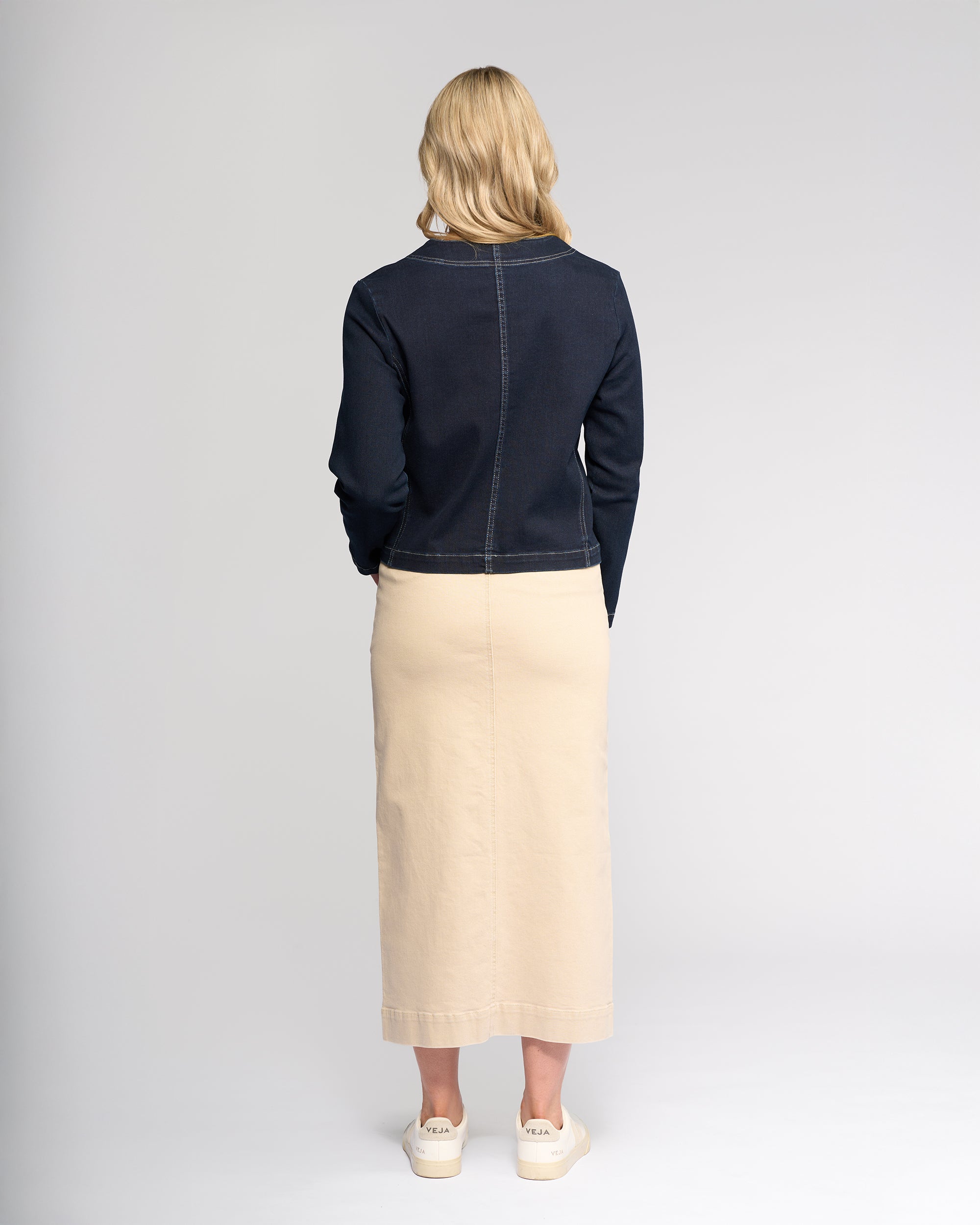 A woman with blonde hair faces away, wearing the Vassalli Cable Jacket - Ink Denim, paired with a beige ankle-length skirt and white sneakers, set against a plain light gray background.