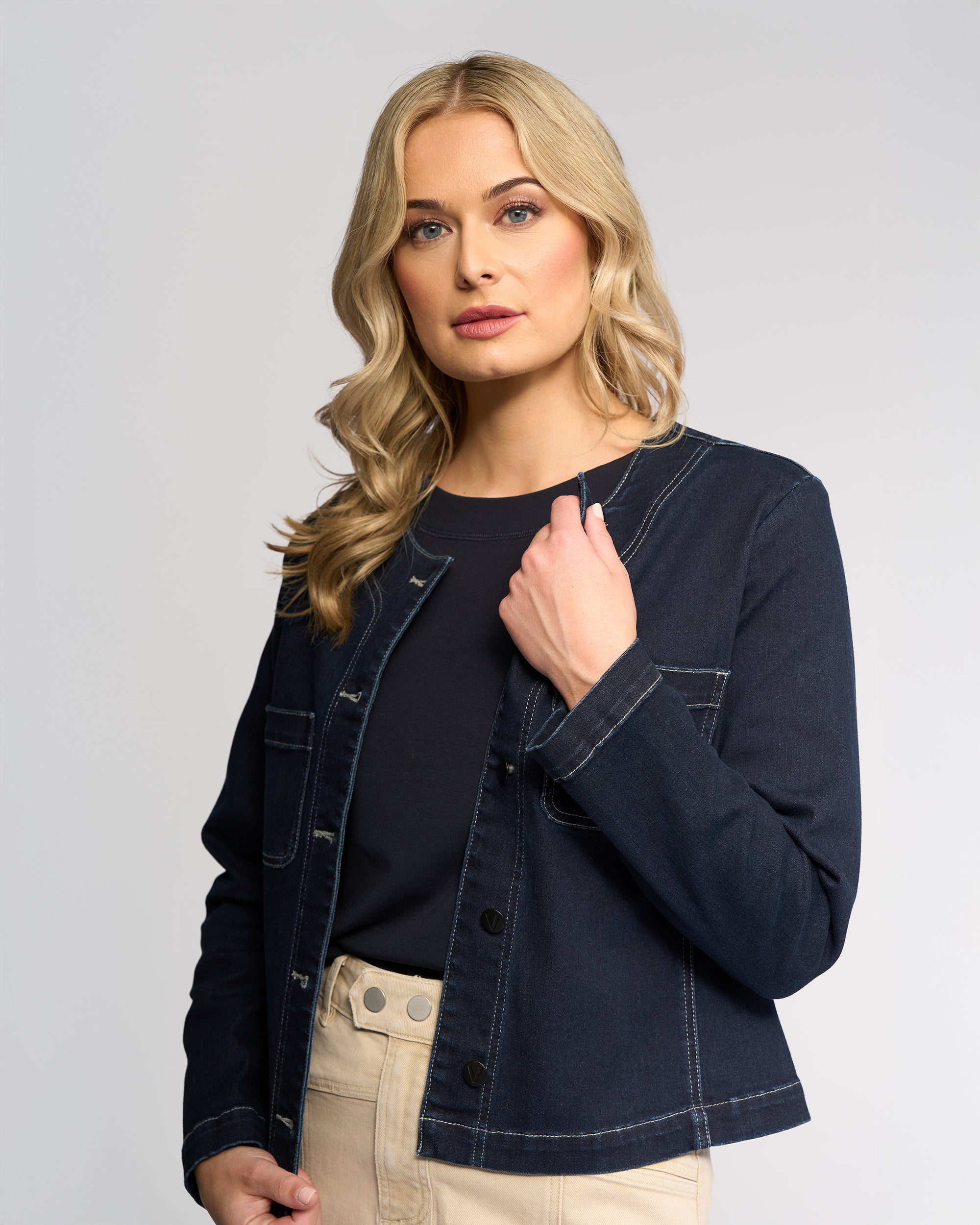 A woman with long blonde hair poses against a plain light background wearing the Vassalli Cable Jacket in Ink Denim over a black top and beige pants, looking at the camera neutrally while holding one lapel.