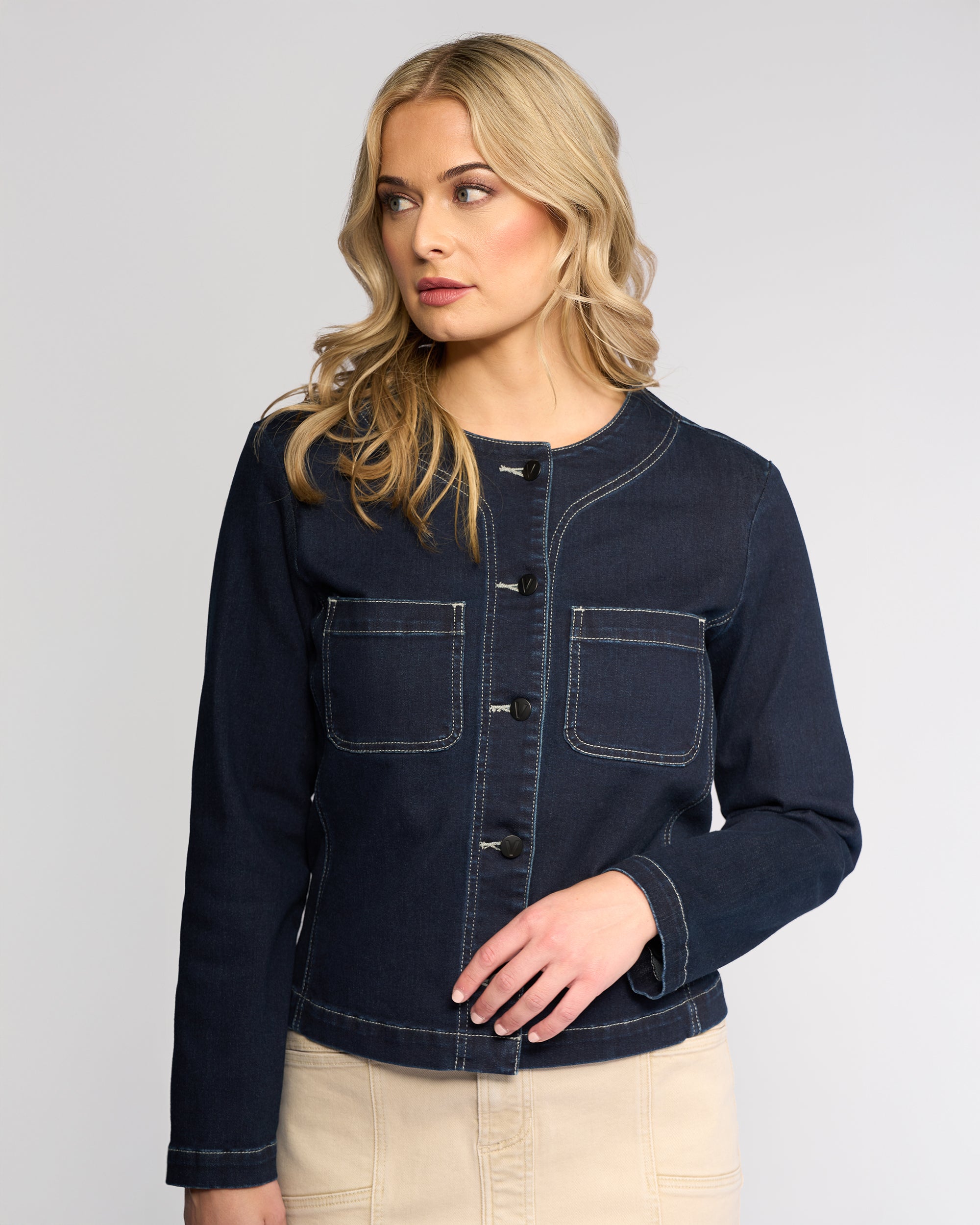 A woman with wavy blonde hair wears the Vassalli Cable Jacket - Ink Denim with front pockets and beige pants, standing against a light grey background and looking slightly to her left.