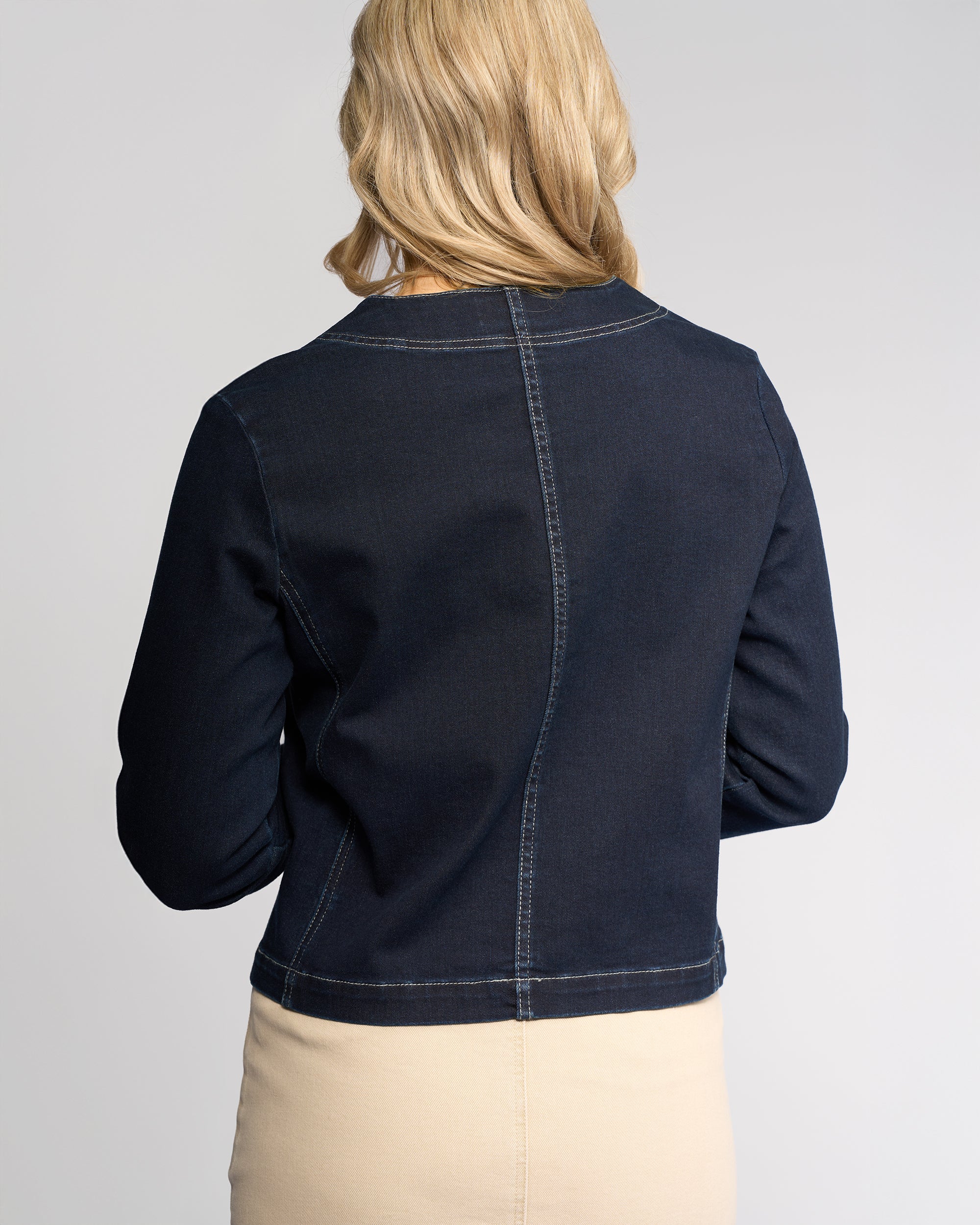 A woman with blonde hair, seen from behind, wears the Vassalli Cable Jacket - Ink Denim in a relaxed fit with a beige skirt. The background is plain and light-colored.