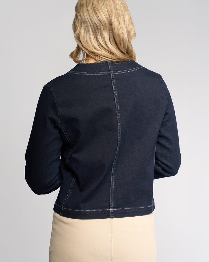 A woman with blonde hair, seen from behind, wears the Vassalli Cable Jacket - Ink Denim in a relaxed fit with a beige skirt. The background is plain and light-colored.
