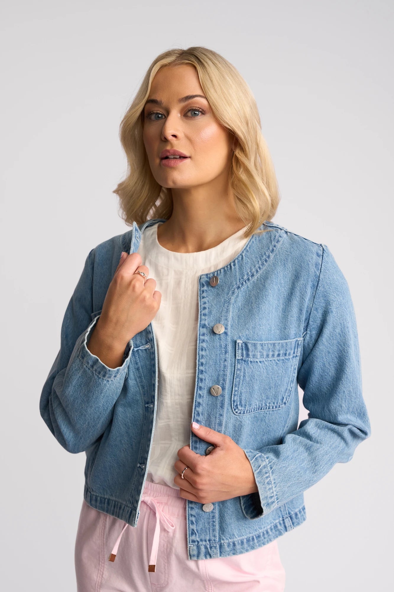 A woman with blonde hair is wearing the Vassalli Cable Jacket in light wash denim over a white top and light pink pants, standing against a plain light background and looking at the camera with a neutral expression.