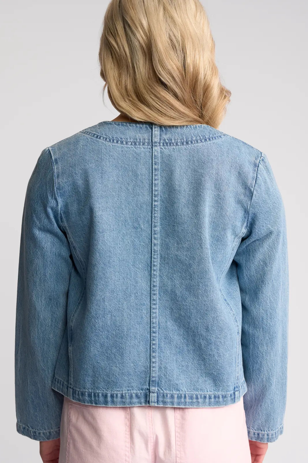 A person with blonde hair is seen from behind, standing against a plain background and wearing the Vassalli Cable Jacket in light wash denim layered over a light pink skirt.