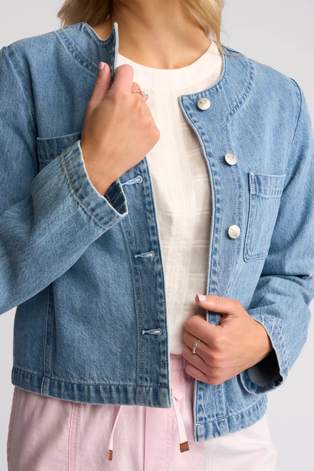 Wearing the Vassalli Cable Jacket in Light Wash Denim over a white shirt and light pink pants, this person touches the jacket collar with one hand while the other hand holds the jacket near the buttons.