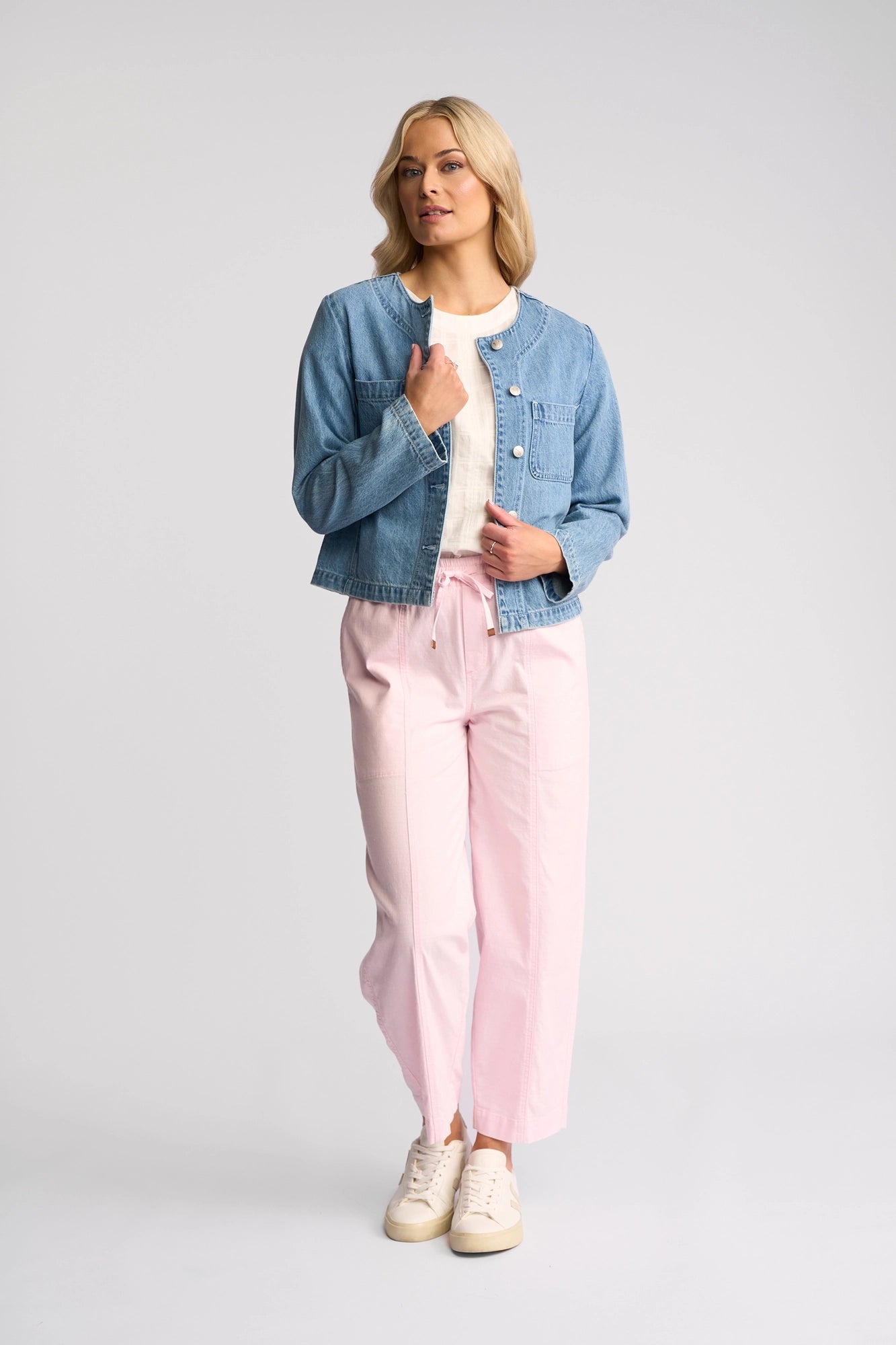 A woman wearing the Vassalli Cable Jacket in light wash denim over a white top, paired with pink wide-leg pants and white sneakers, stands against a plain light background, gazing slightly to the side with one hand touching her jacket.