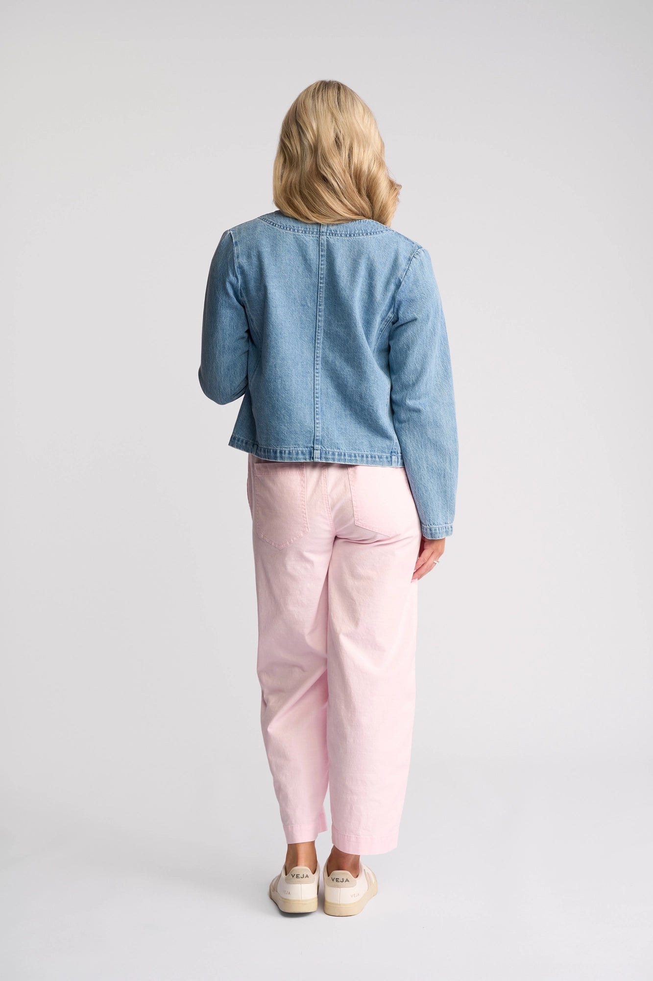 A person with blonde hair stands facing away, wearing the Vassalli Cable Jacket in light wash denim, paired with pale pink pants and white sneakers, against a plain white background.