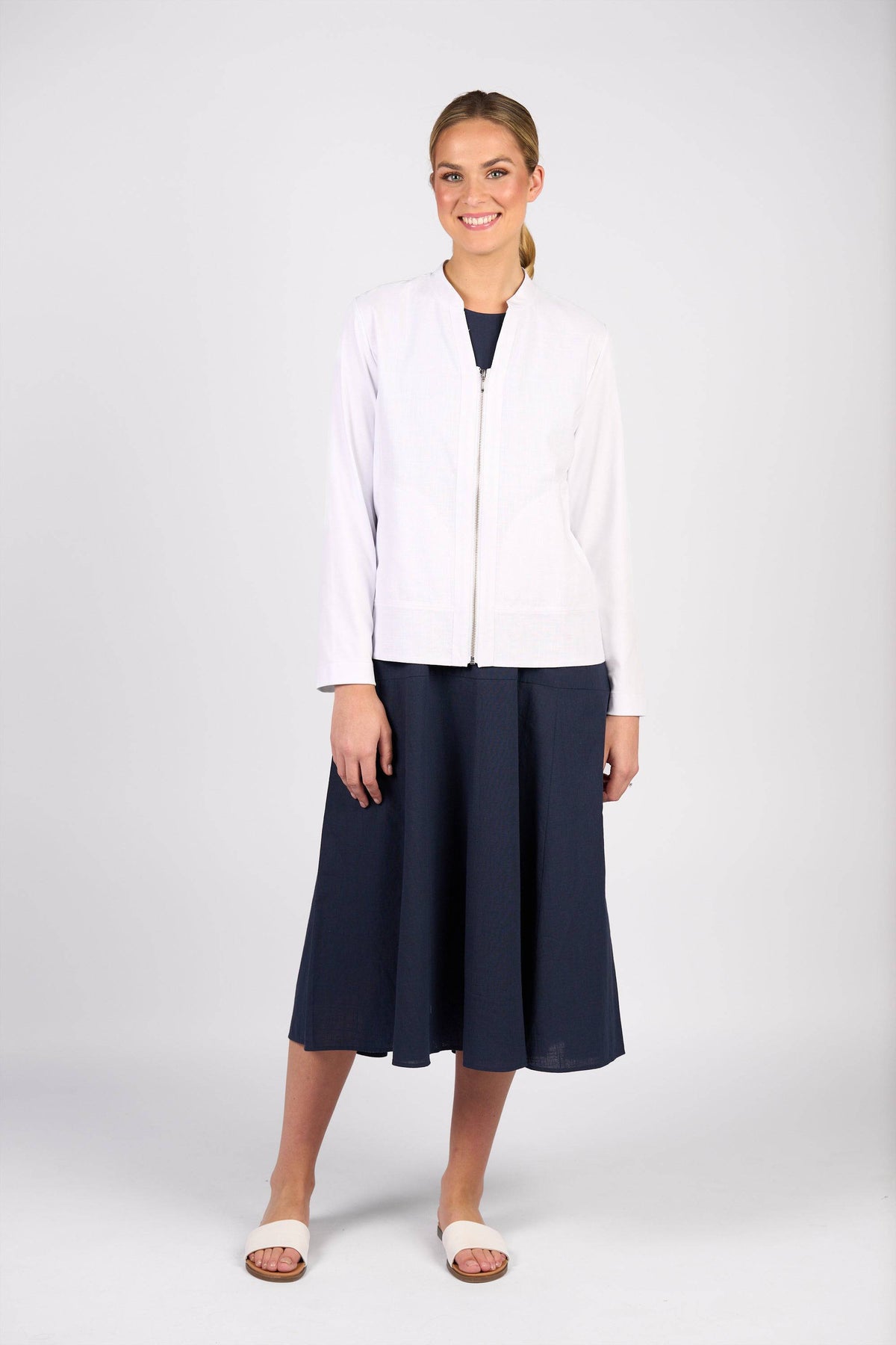 A woman smiles against a plain background, wearing the Vassalli Zip Up Jacket in white over a knee-length navy dress and white sandals.