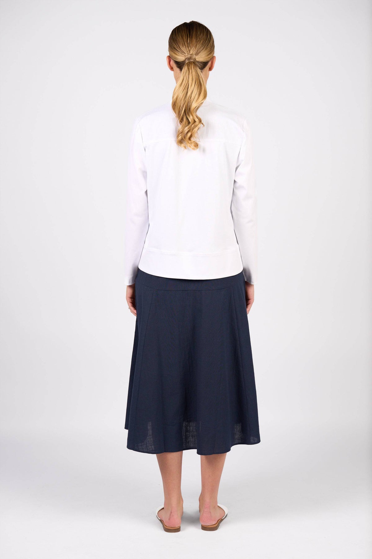 A woman with blonde hair in a ponytail faces away, wearing the Vassalli Zip Up Jacket in white, a dark blue midi skirt, and white slip-on shoes against a plain white background.