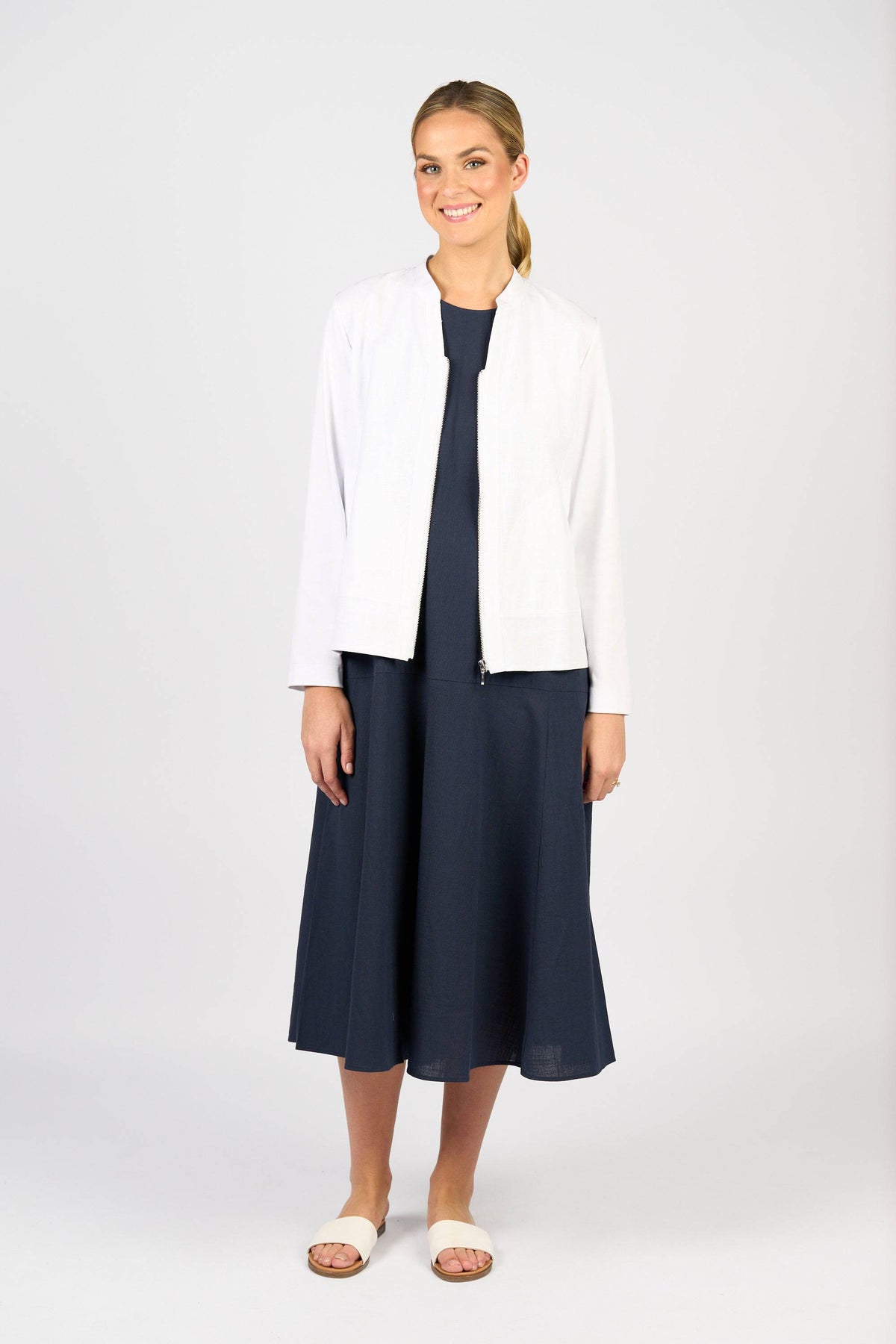 A woman stands smiling against a plain background, wearing the Vassalli Zip Up Jacket in white over a navy blue midi dress with light-colored sandals. Her hair is tied back in a low ponytail.