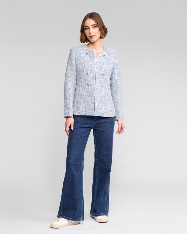 A woman with shoulder-length hair and a neutral expression stands against a plain background, wearing the Vassalli Lena Jacket - Valli, blue flared jeans, and white platform shoes.