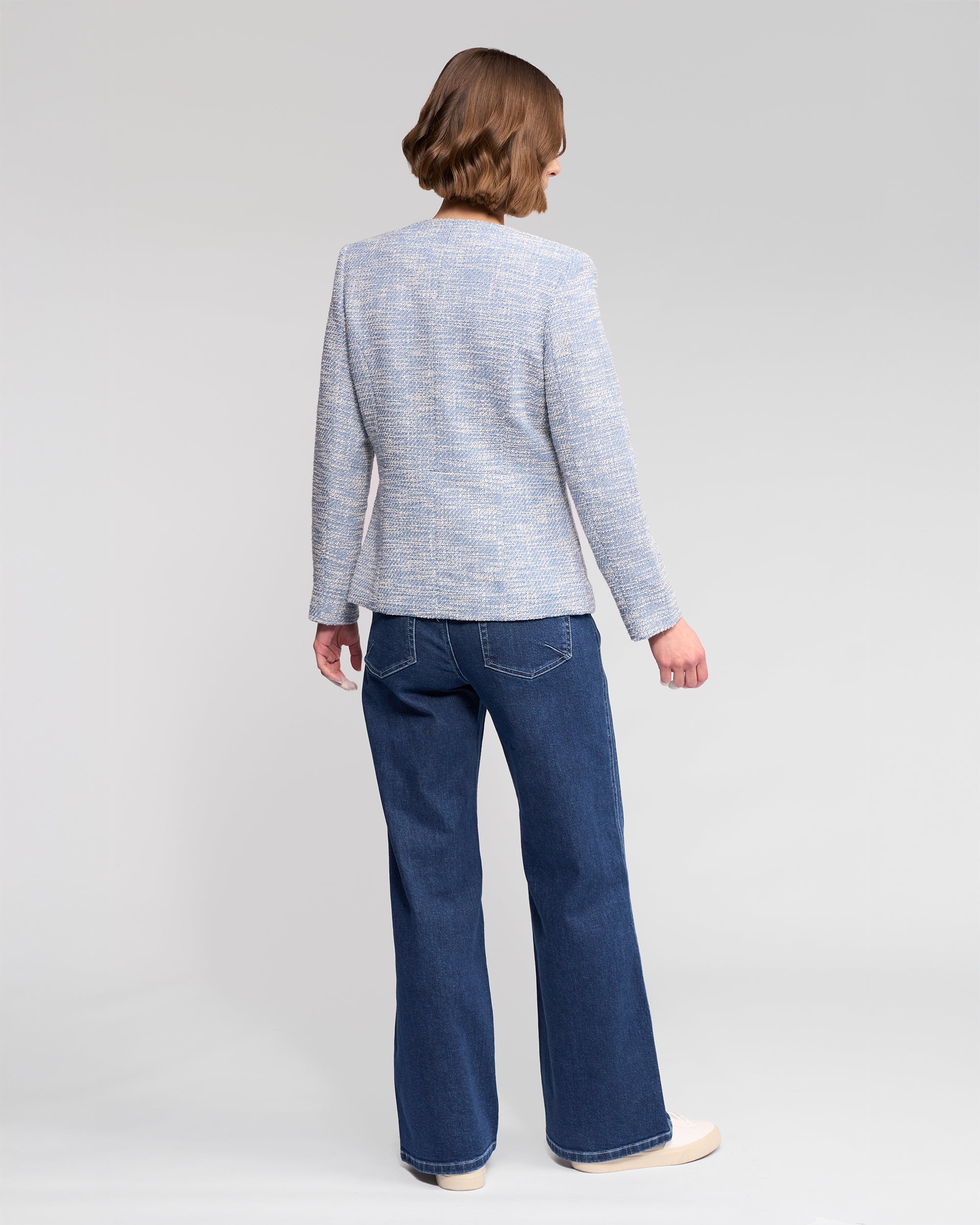 A person with short brown hair, dressed in a Vassalli Lena Jacket - Valli in light gray, dark blue jeans, and beige shoes, stands facing away from the camera against a plain light background.