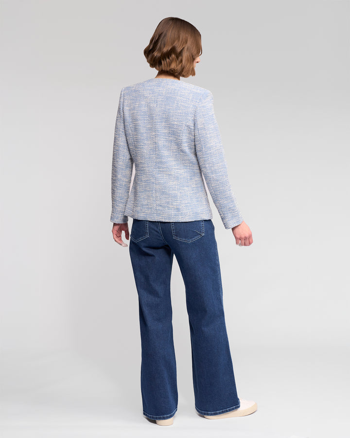 A person with short brown hair, dressed in a Vassalli Lena Jacket - Valli in light gray, dark blue jeans, and beige shoes, stands facing away from the camera against a plain light background.