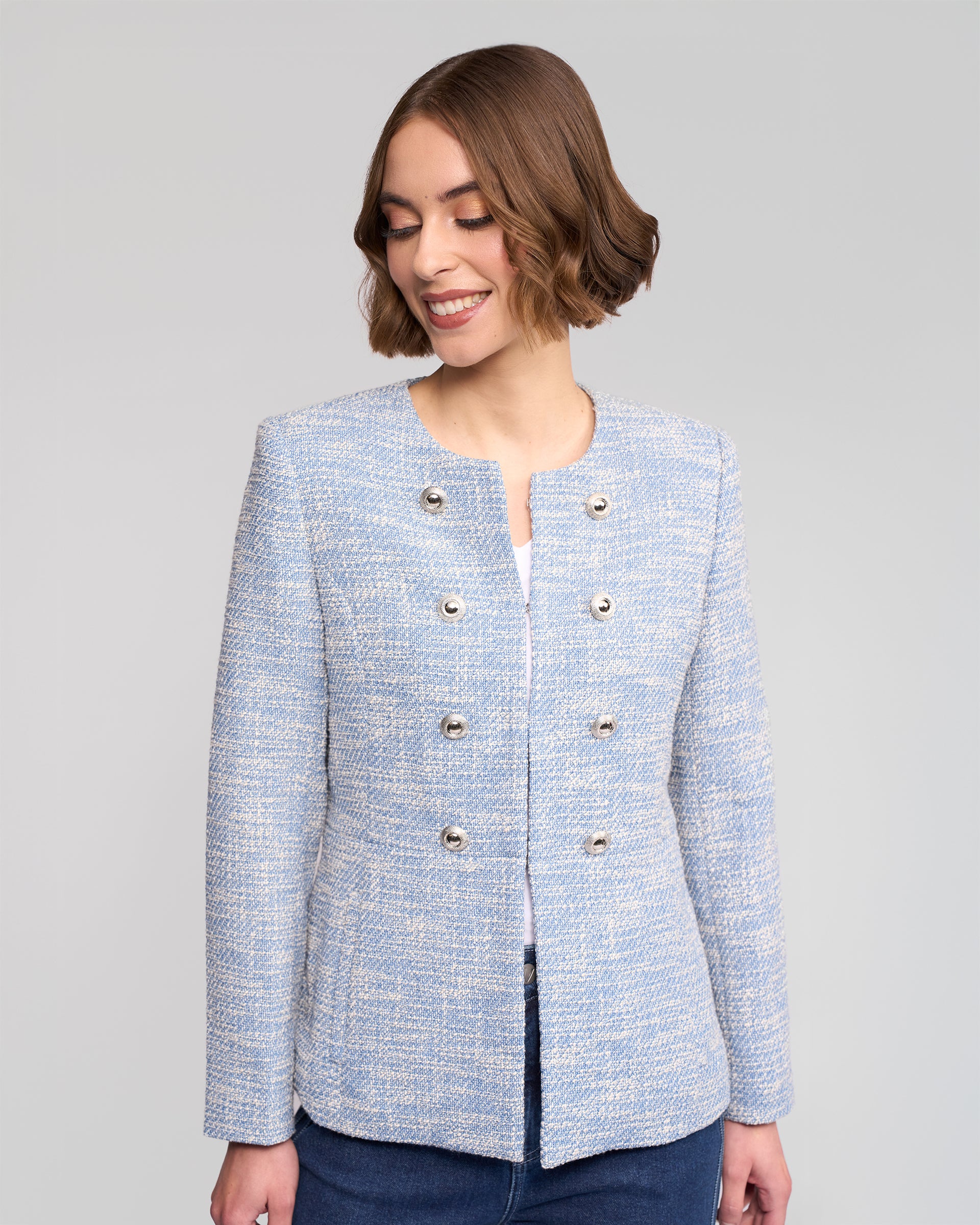 A woman with short brown hair smiles while wearing the Lena Jacket - Valli by Vassalli, a light blue woven jacket with silver buttons styled over a white top and blue jeans, posed against a plain light gray background.