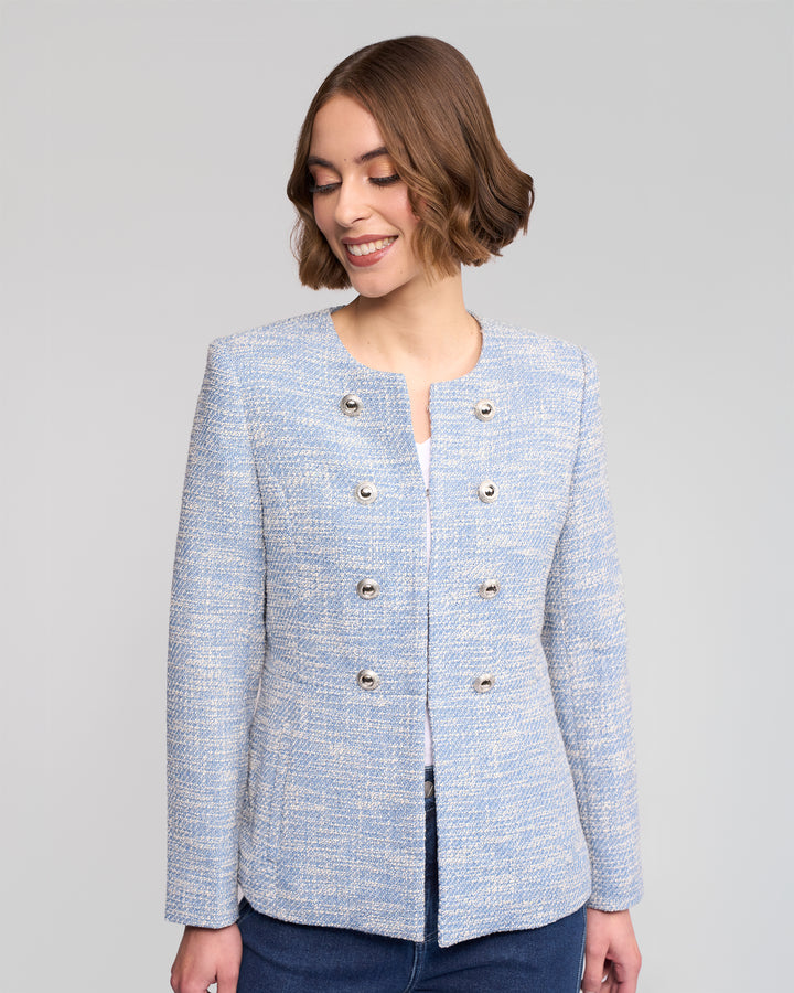 A woman with short brown hair smiles while wearing the Lena Jacket - Valli by Vassalli, a light blue woven jacket with silver buttons styled over a white top and blue jeans, posed against a plain light gray background.