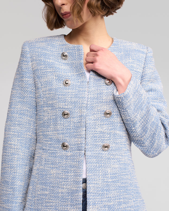 Woman wears the Vassalli Lena Jacket - Valli, a textured light blue woven jacket with metallic buttons, layered over a white top. Her wavy chin-length hair frames her face, which is partially out of frame.