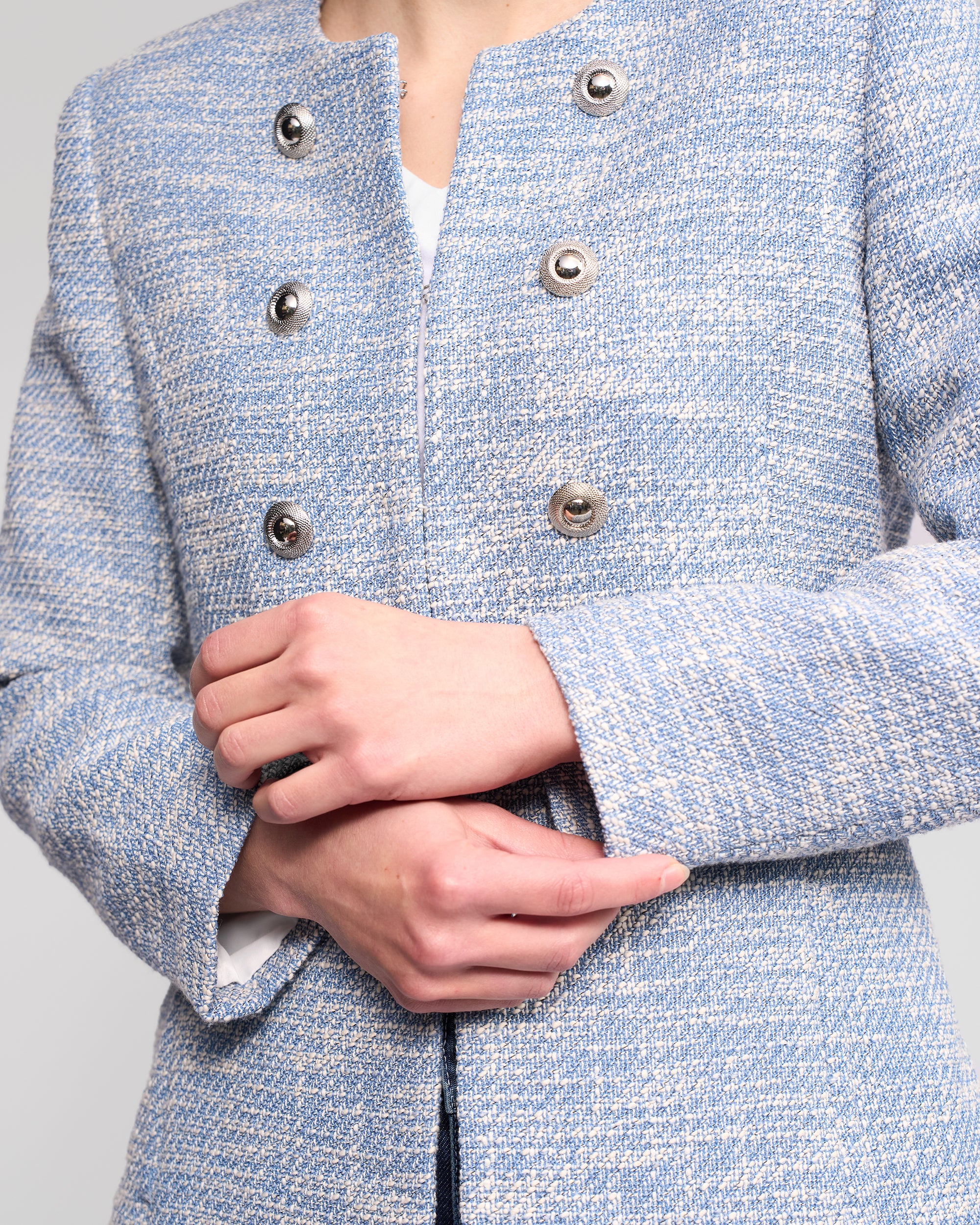 A person wearing the Lena Jacket - Valli by Vassalli, a light blue woven women’s jacket with metallic buttons and no collar, adjusts the sleeve. Its structured, stylish design makes it perfect for formal occasions.