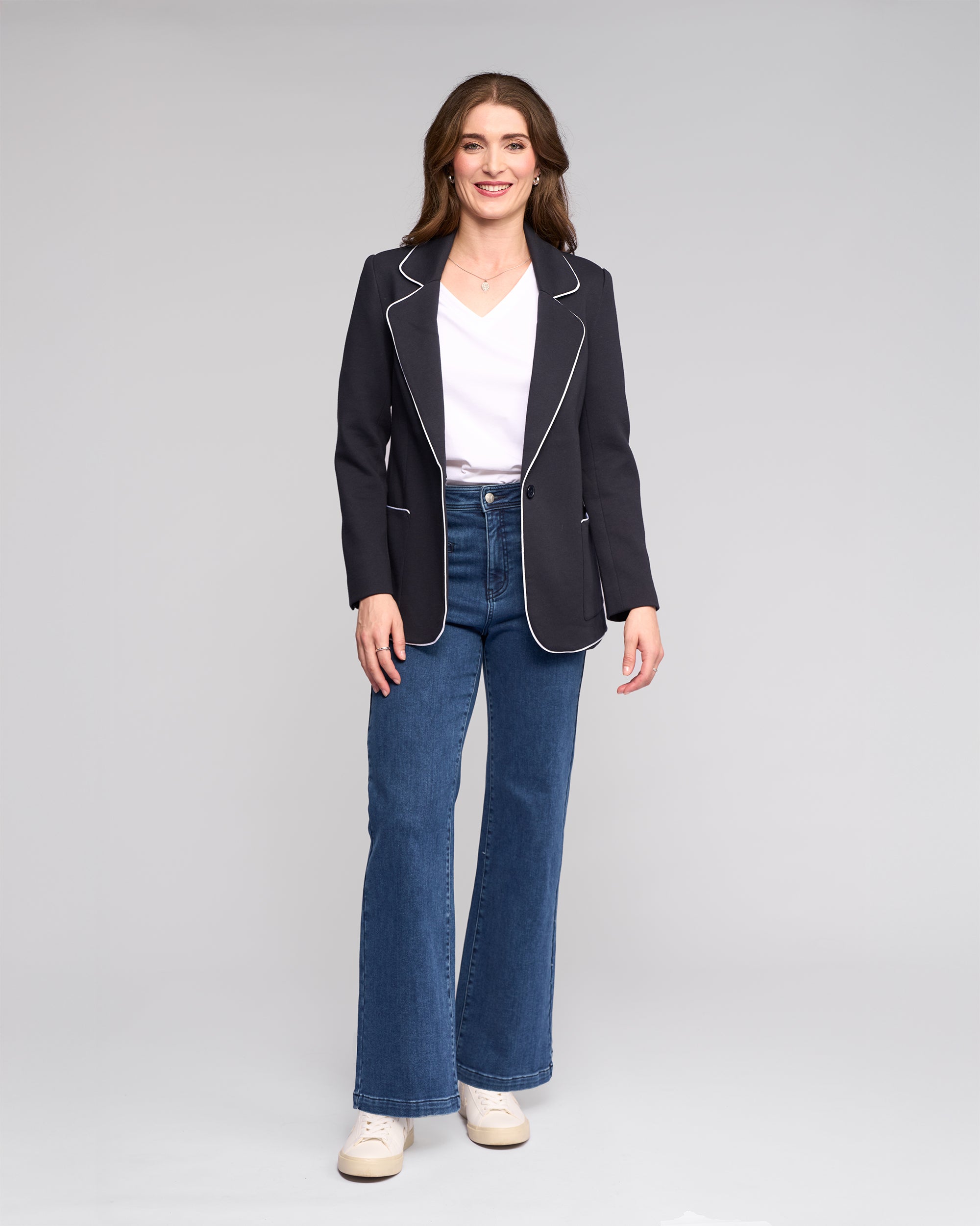A woman stands and smiles, wearing the Vassalli Ora Blazer - Ink, a white V-neck top, blue flared jeans, and white sneakers against a plain light gray background.